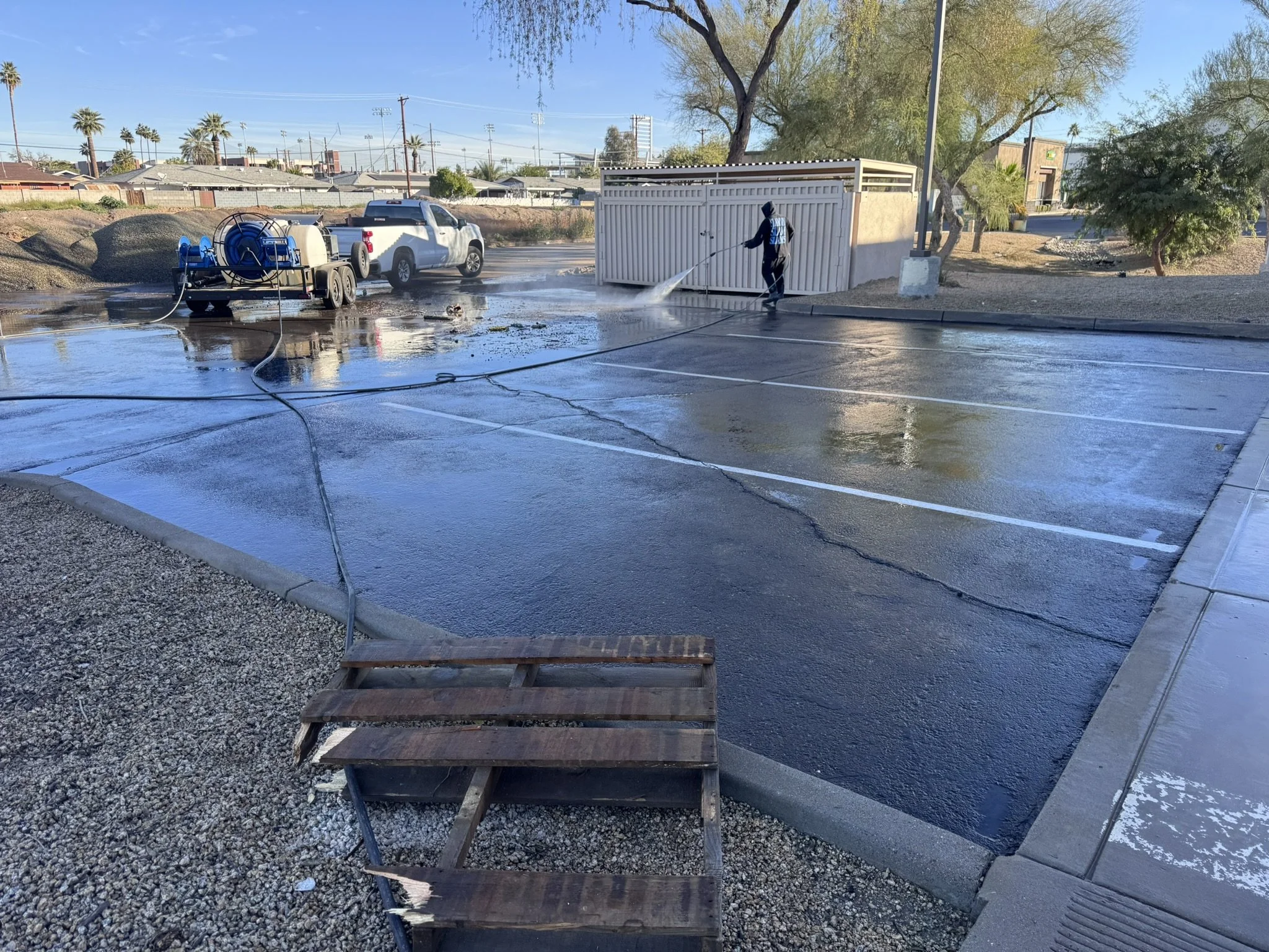 A person using a pressure washer to clean a parking lot surface, with a trailer and hose connected nearby. Trees and cars are visible in the background on a sunny day.