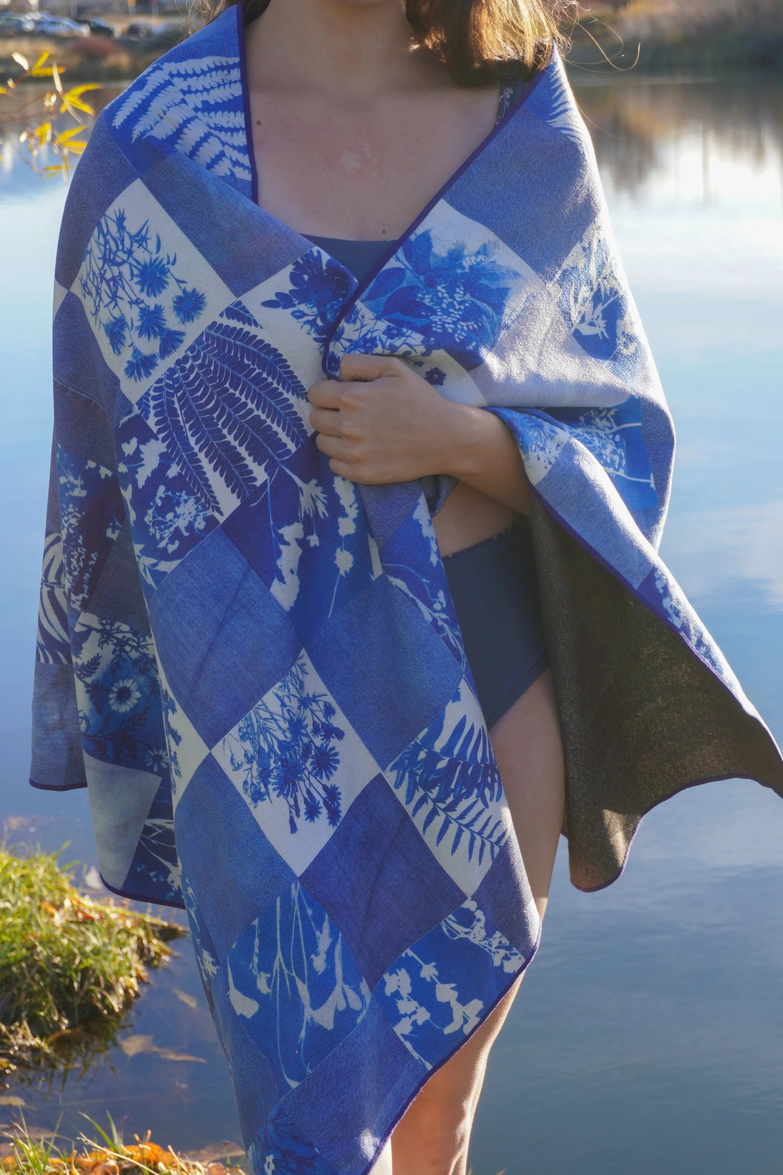 Person wrapped in a blue and white floral patterned towel standing near a body of water during sunset.