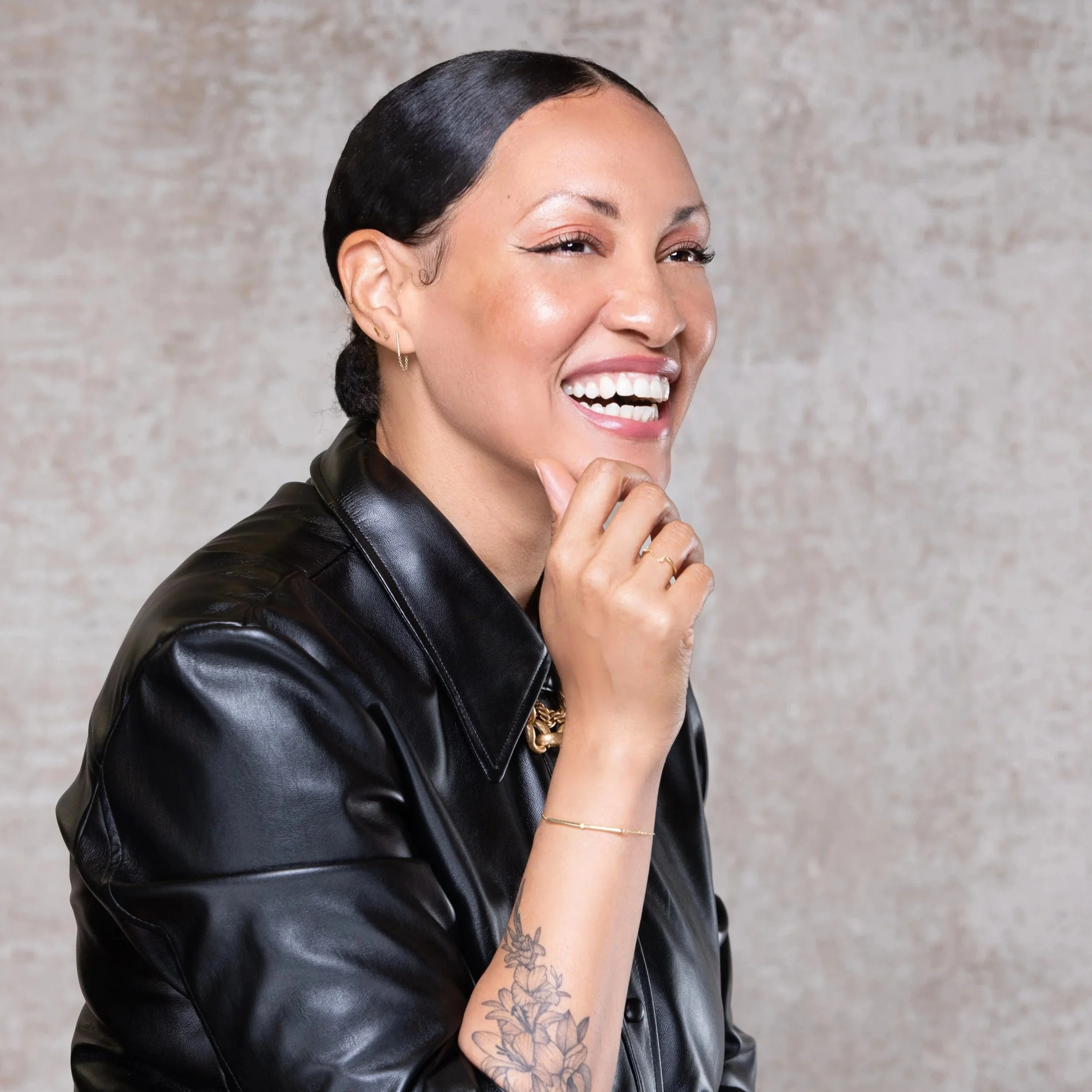 A woman with slicked-back black hair, dressed in a black leather jacket with a gold chain necklace, standing in front of green plant leaves.