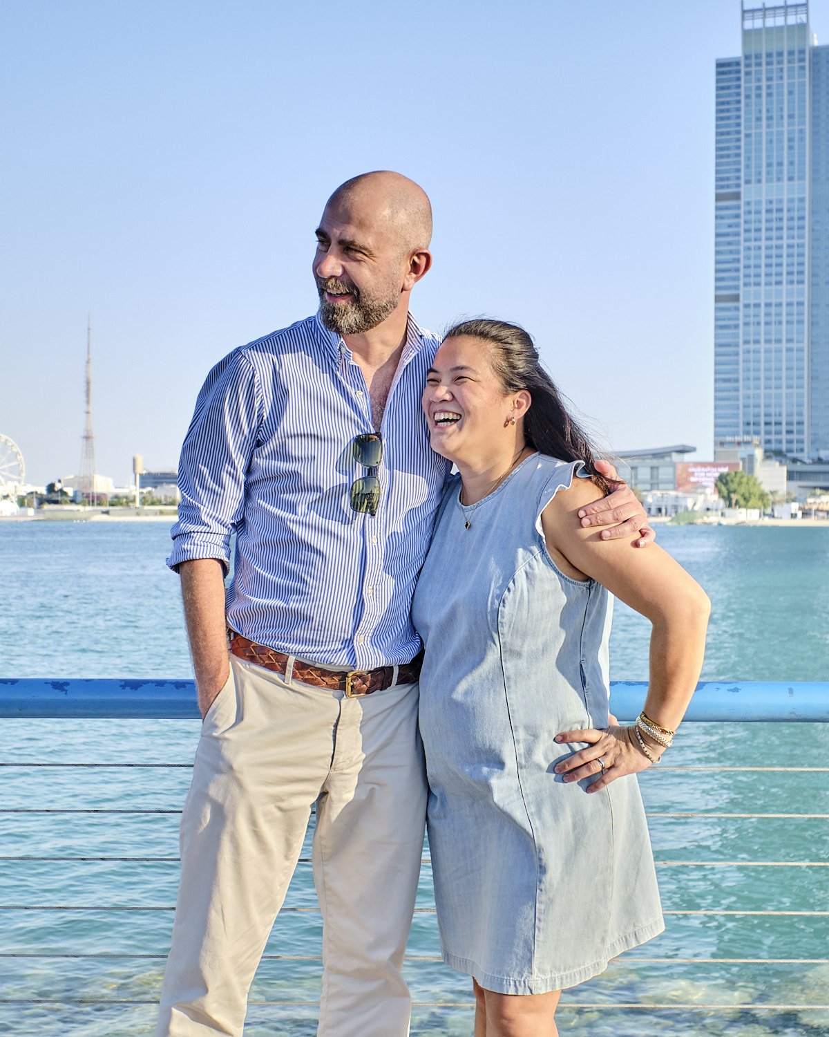 A couple smiling by a waterfront with modern buildings in the background.