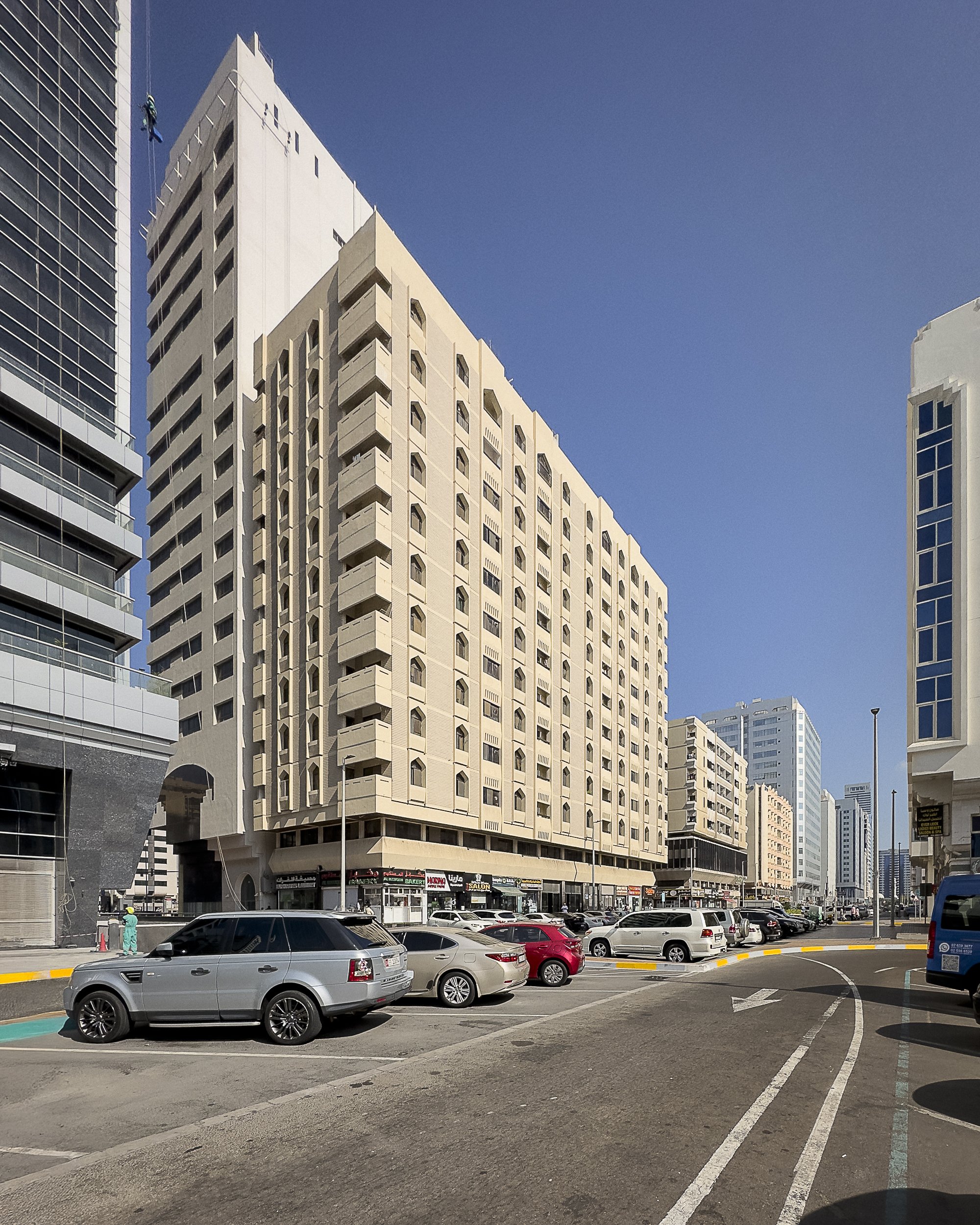Urban street scene with tall beige building, parked cars, and clear blue sky.