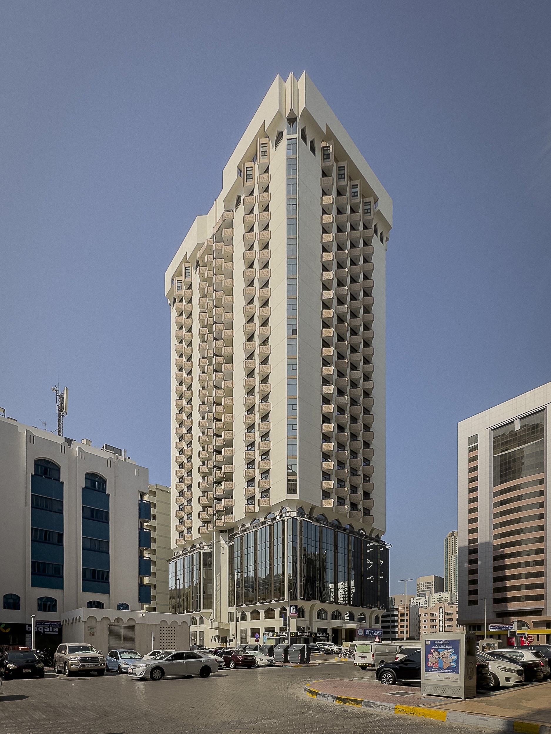 Tall modern building with balconies in an urban area, surrounded by smaller buildings and parked cars under a clear sky.