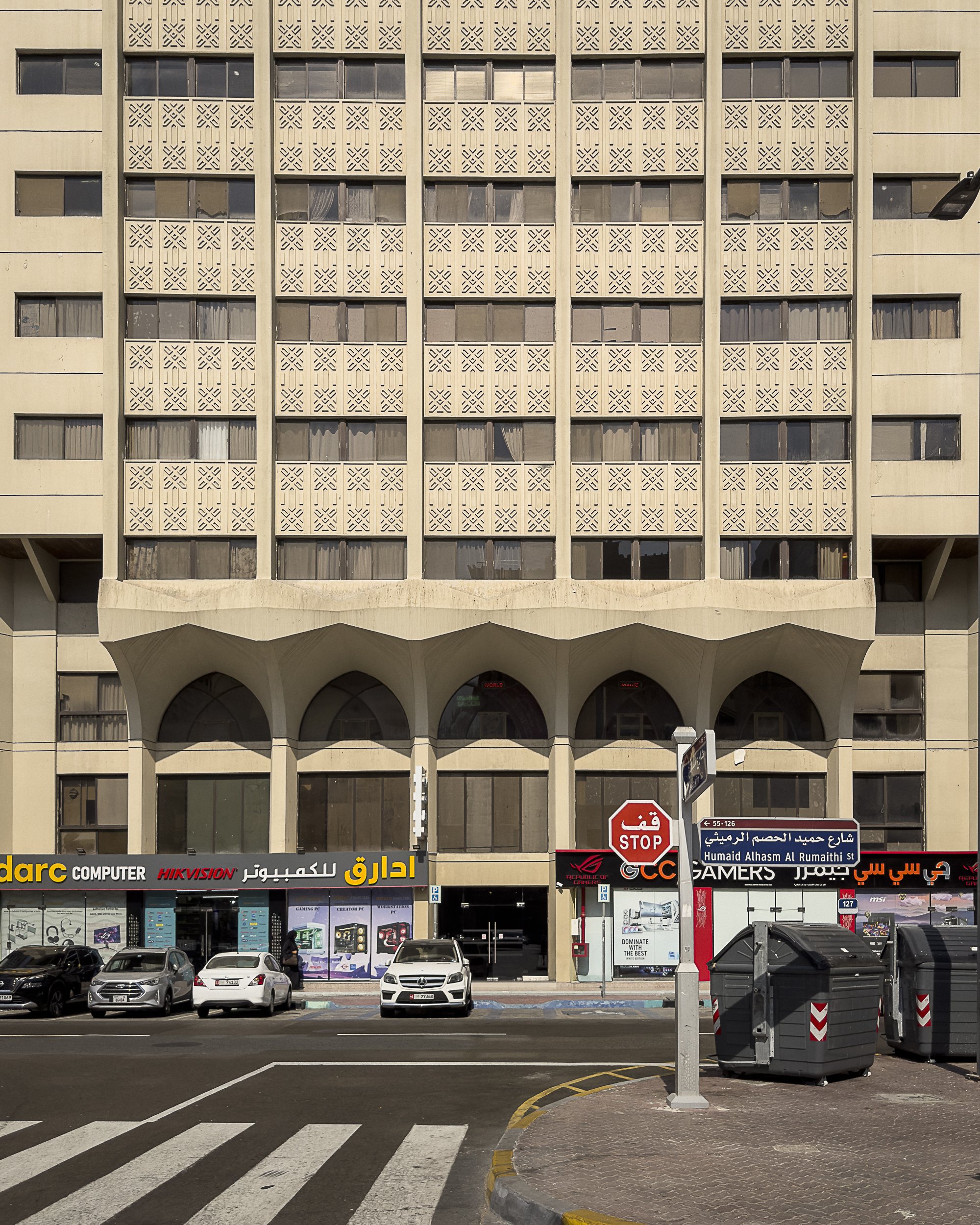 Building facade with geometric patterns, shops, cars parked, and stop sign on street corner in urban setting.