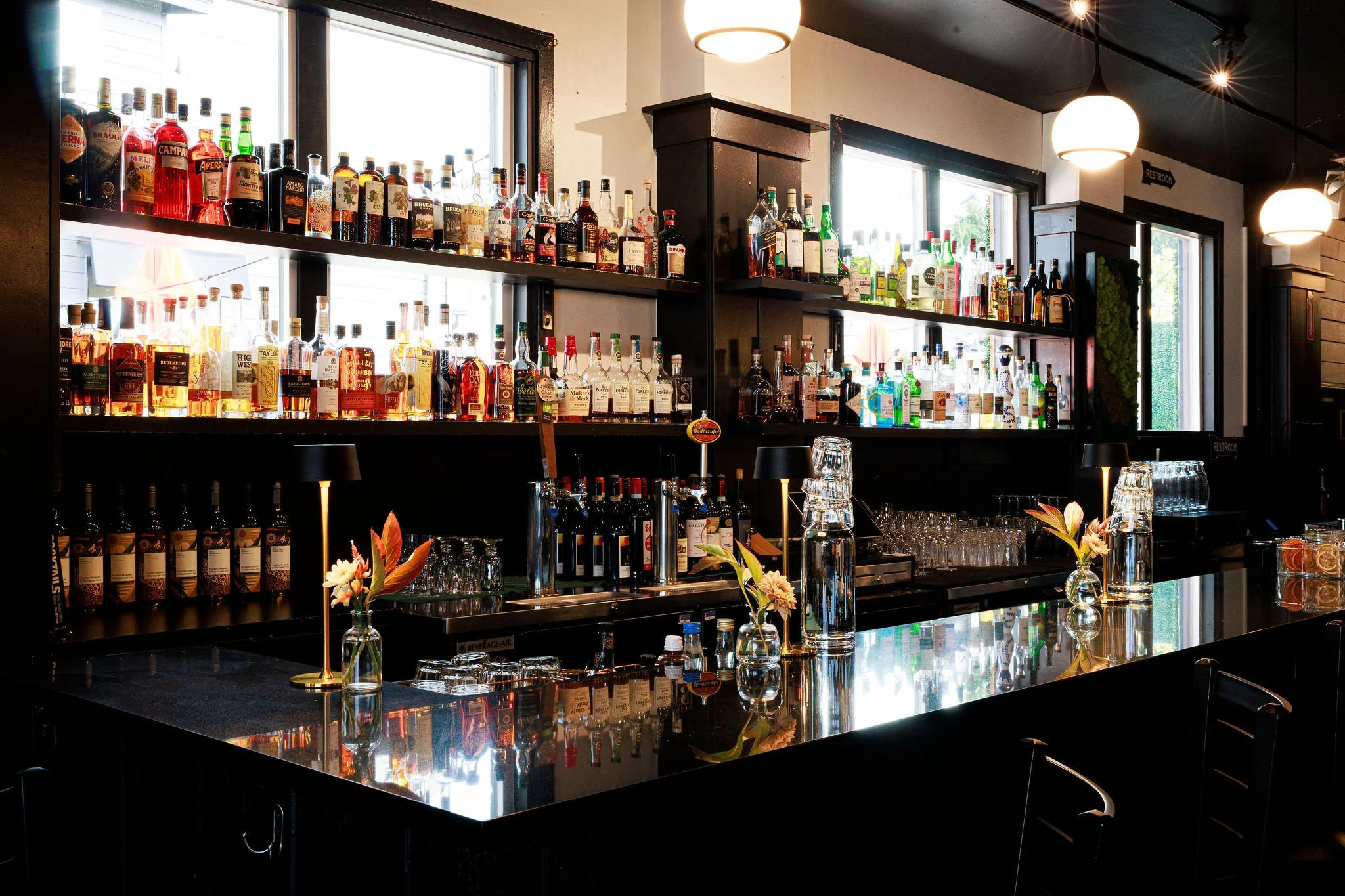 A bar with alcohol bottles on illuminated shelves behind a black counter, decorated with small vases with flowers, in front of windows.