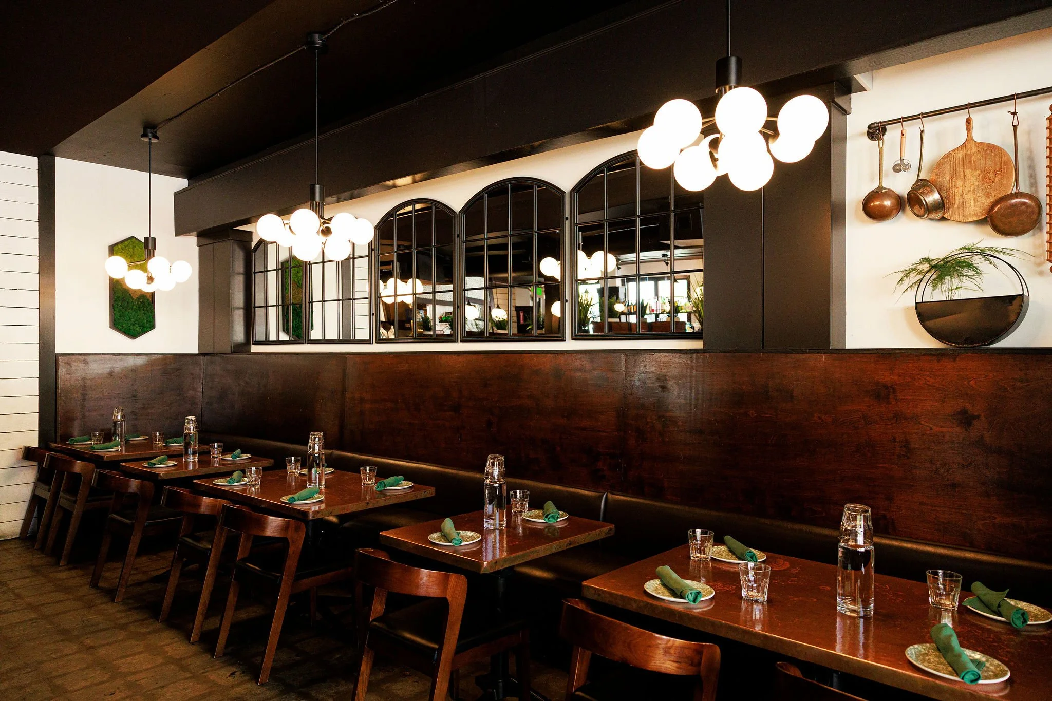 Interior of a restaurant with wooden tables, green napkins, water glasses, and dark wooden chairs, decorated with modern hanging light fixtures and hanging rustic kitchen utensils on the wall.