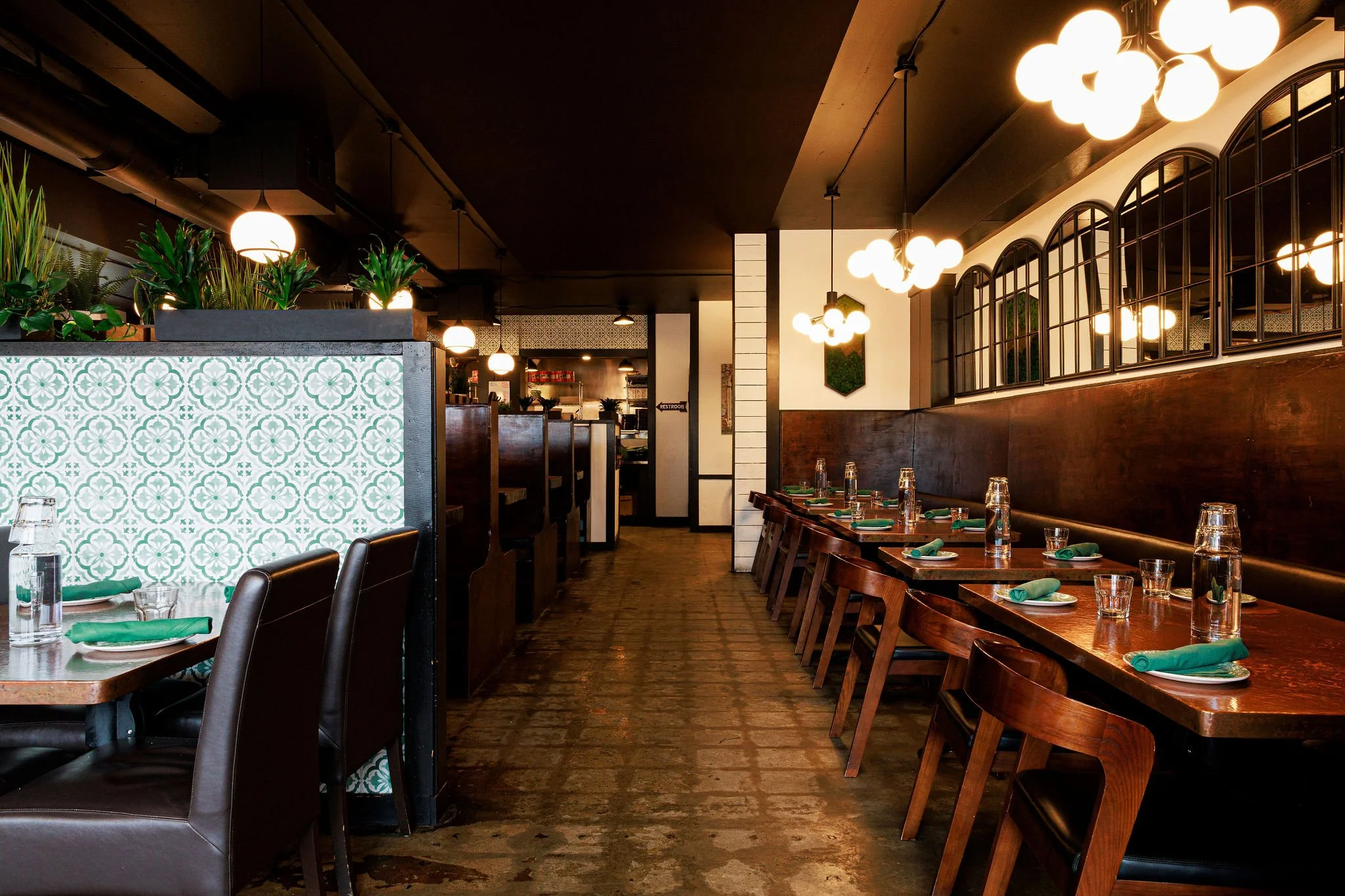 Empty restaurant dining area with wooden tables and chairs, set with water glasses and green napkins, illuminated by modern round light fixtures