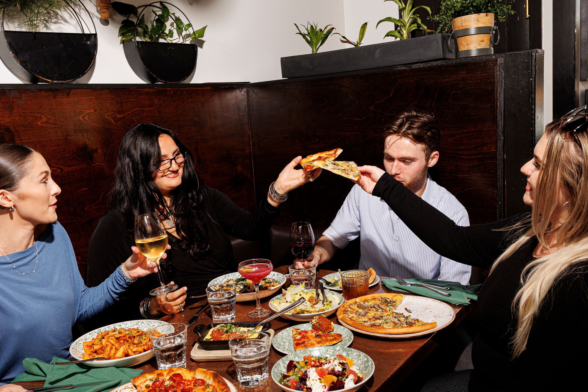 Group of four friends enjoying dinner at a restaurant, sharing pizza, pasta, and salad, with glasses of wine and drinks on the table.