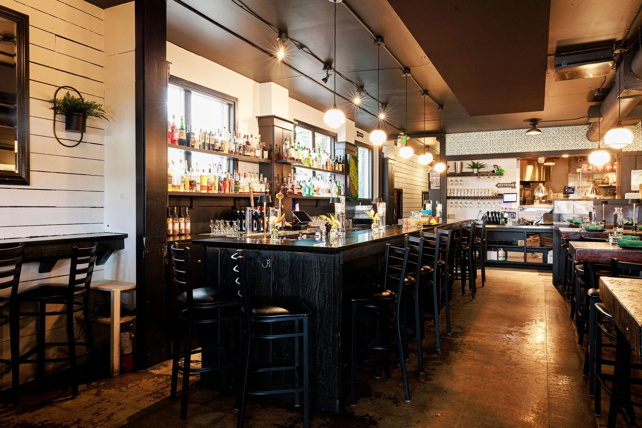 Empty bar inside a restaurant with a black countertop, tall black chairs, and shelves with bottles of liquor. Dim lighting with hanging lights and a mix of modern and rustic decor.