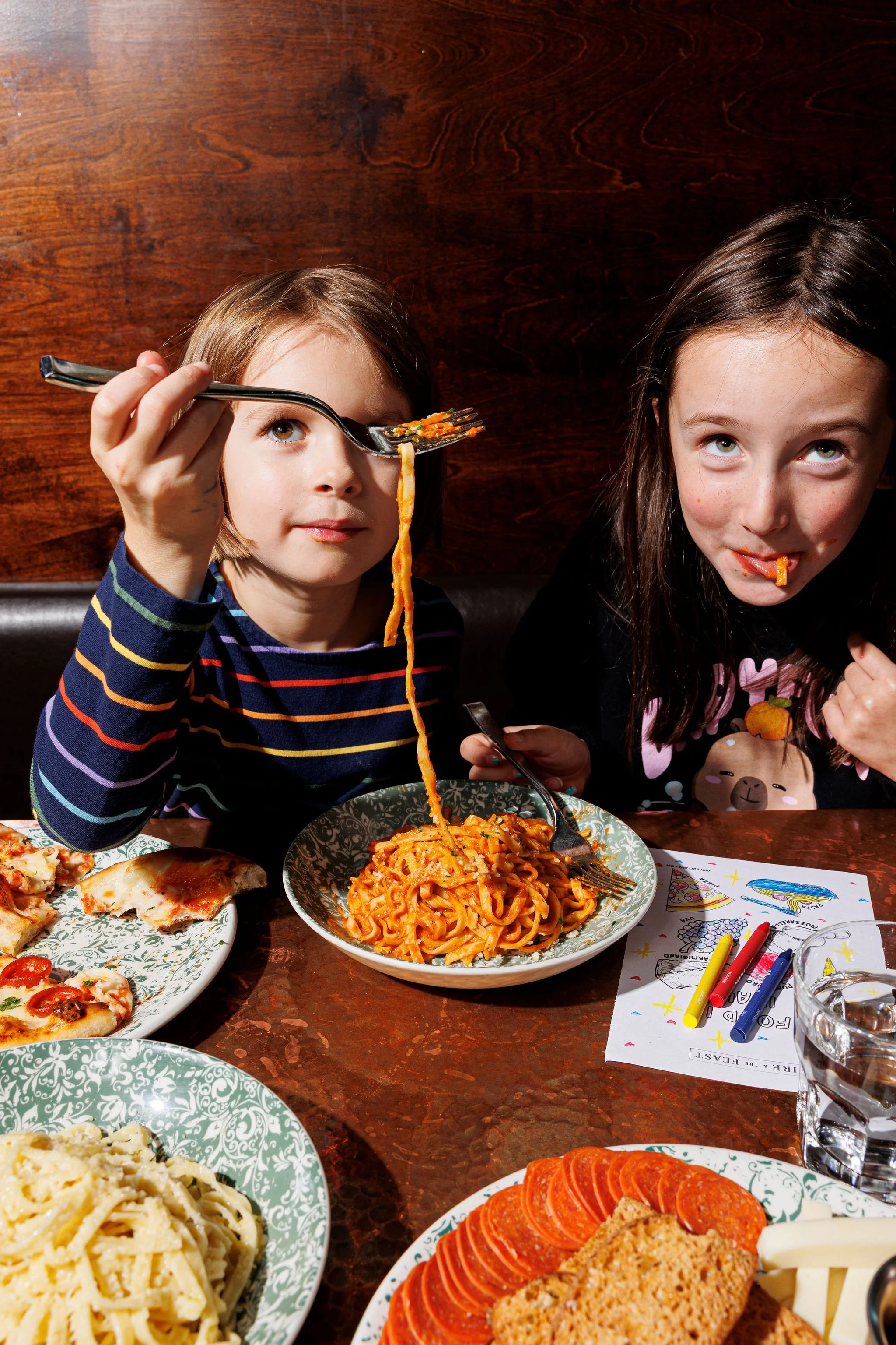 Two young girls eating spaghetti at a dinner table, with plates of pizza, pasta, and sliced meat, and coloring items on the table.