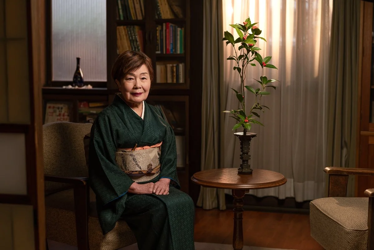 An elderly Japanese woman in a green kimono sitting in a room with a wooden table and a tall plant with red berries.
