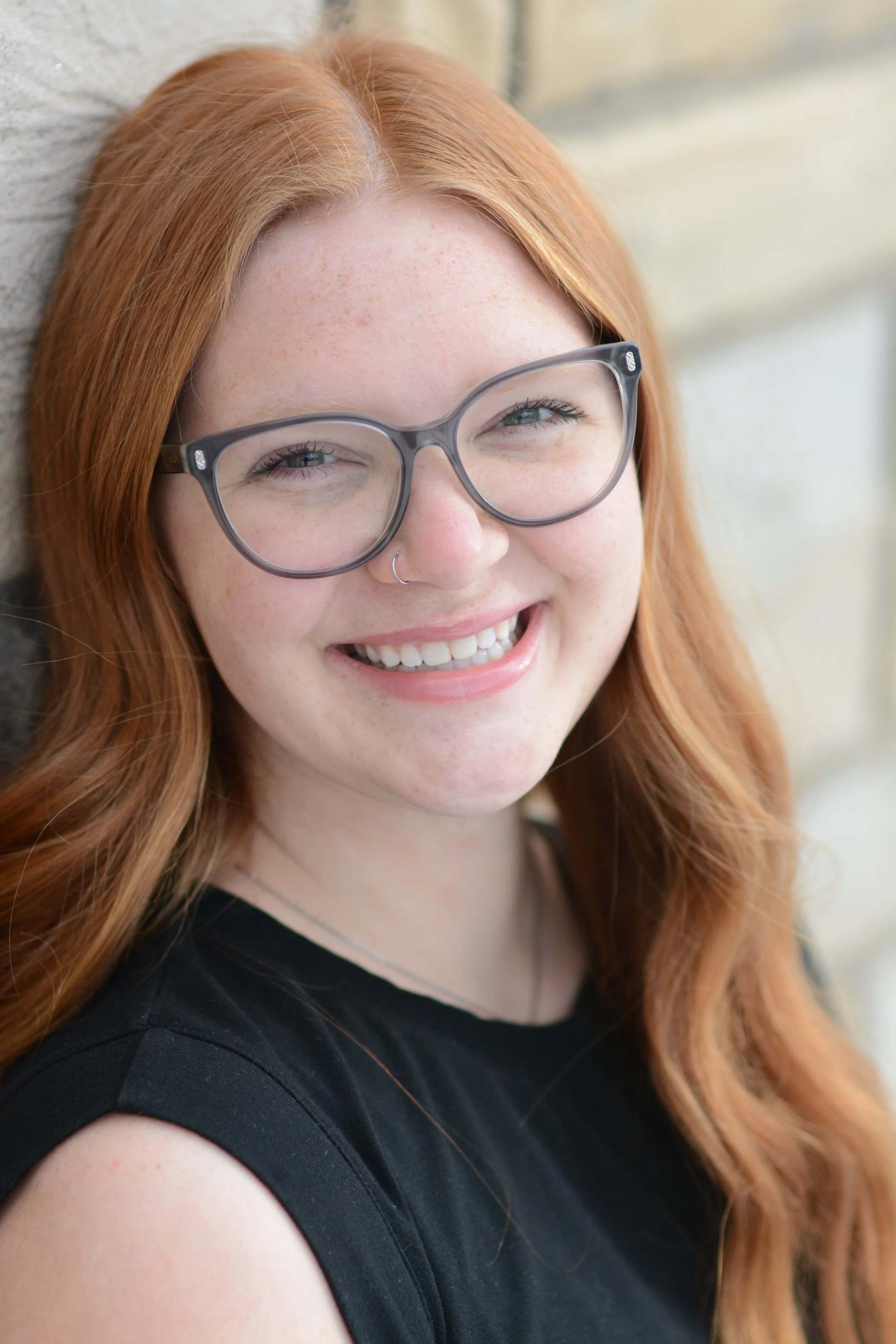 Close-up of a young woman with red hair, glasses, and a nose ring, smiling and looking at the camera, against a blurred outdoor background.