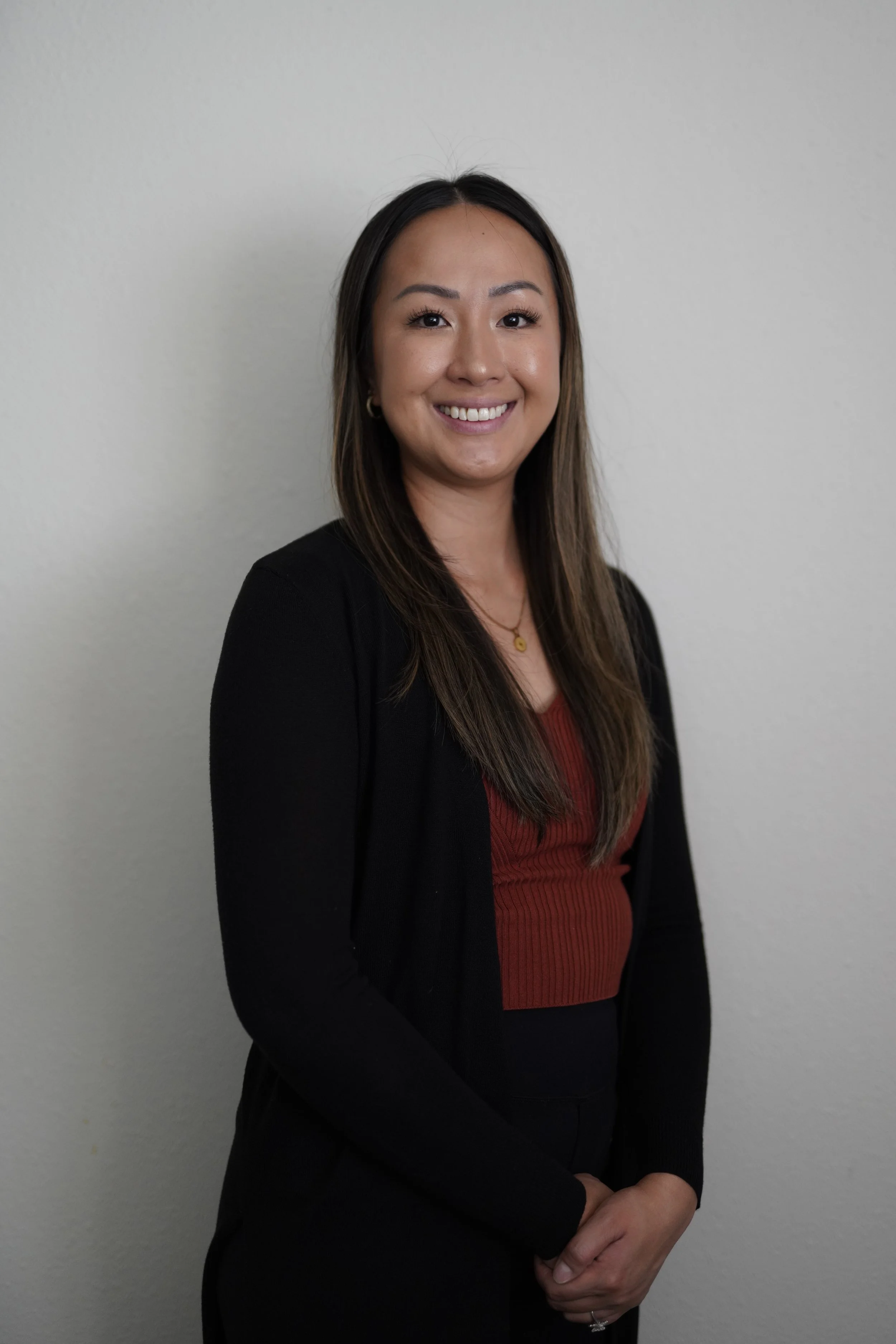 A woman with long dark hair, wearing a black blazer over a red top, standing in front of a plain light gray wall, smiling with her hands clasped in front of her.