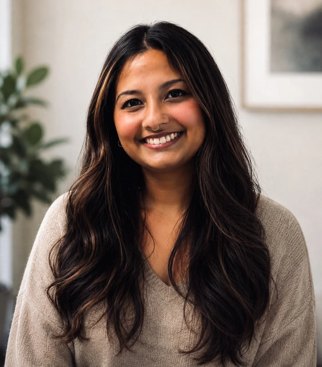 A woman with long dark hair, smiling, wearing a beige sweater, in an indoor setting with a plant and framed picture in the background.