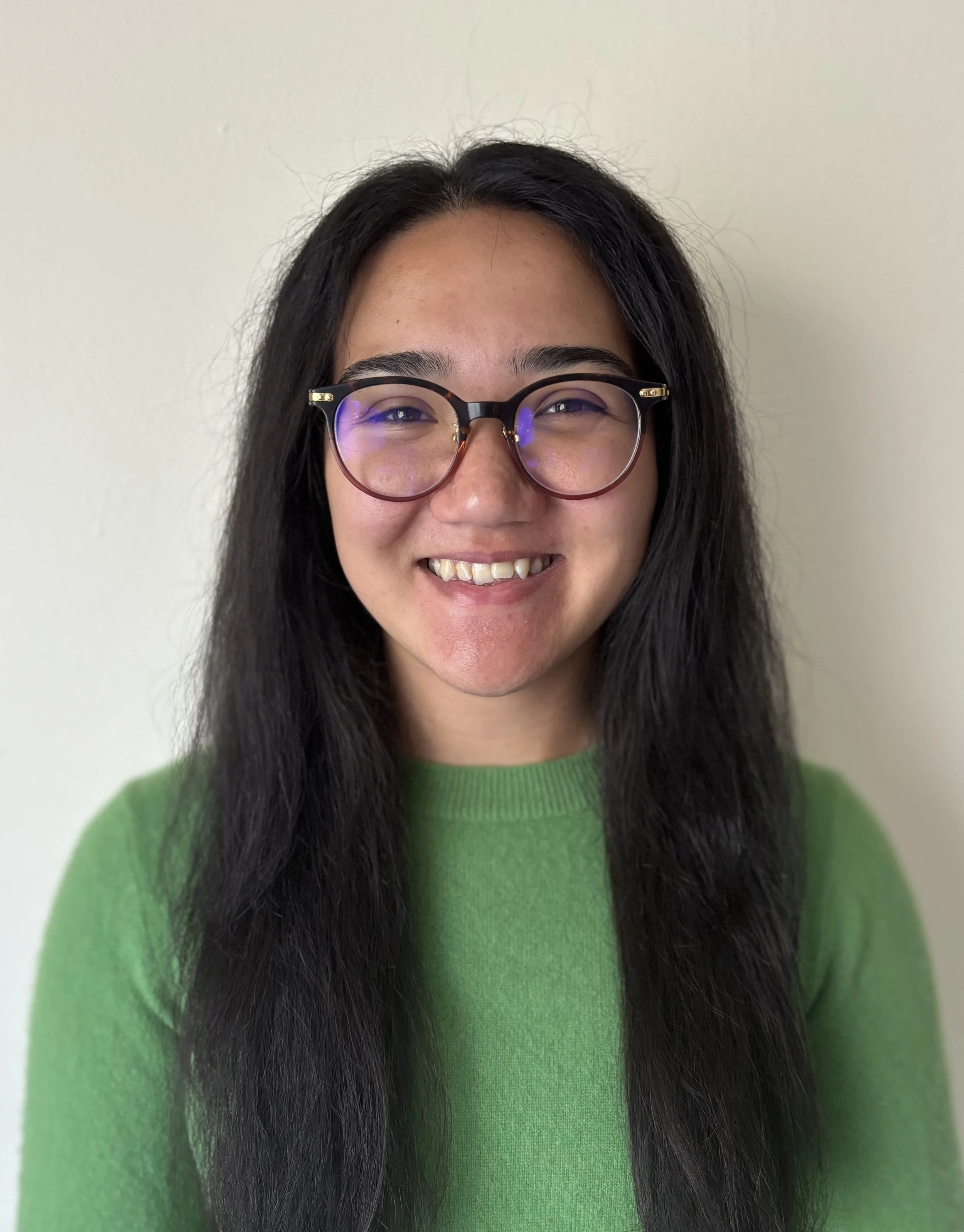 Close-up of a smiling woman with long black hair wearing tortoiseshell glasses and a green sweater, against a plain white wall.