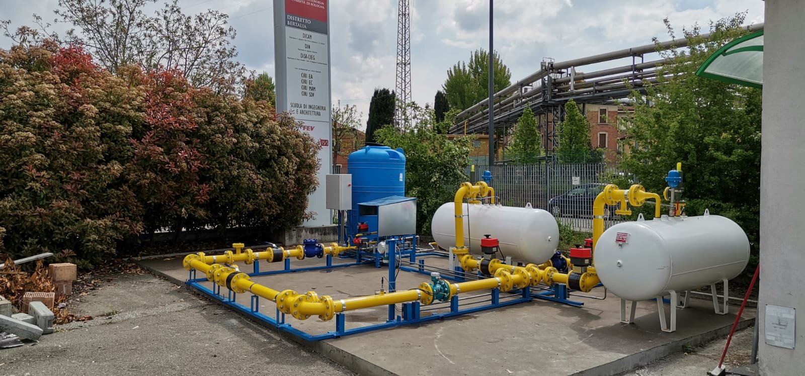 Impianto di trattamento acqua con serbatoi e tubature gialle, situato all'aperto vicino a delle piante e un'area urbana.