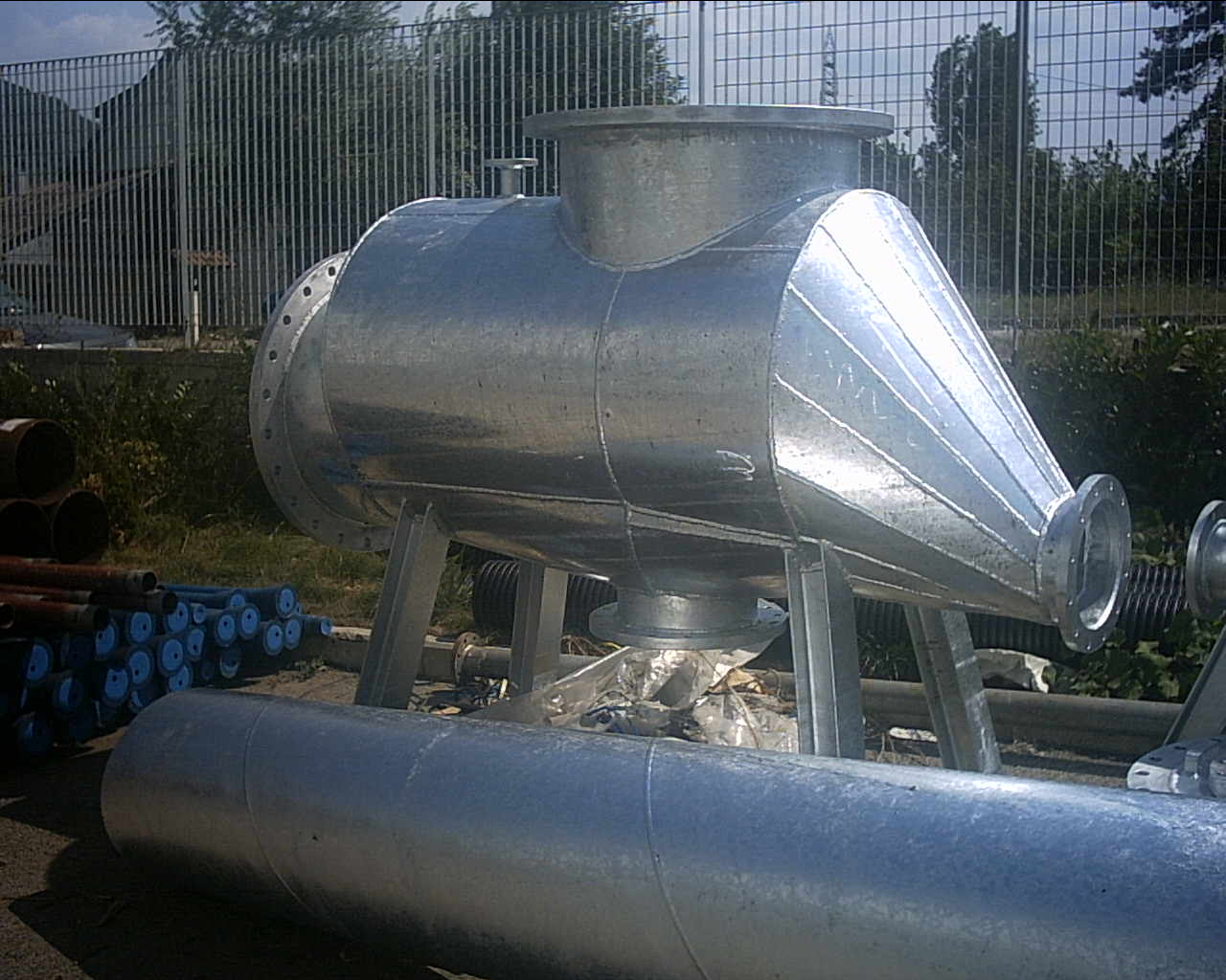 Immagine di una grande camera d'aria di metallo, probabilmente un componente di aspirazione o scarico, posta all'aperto con tubi di metallo di varie dimensioni sullo sfondo.