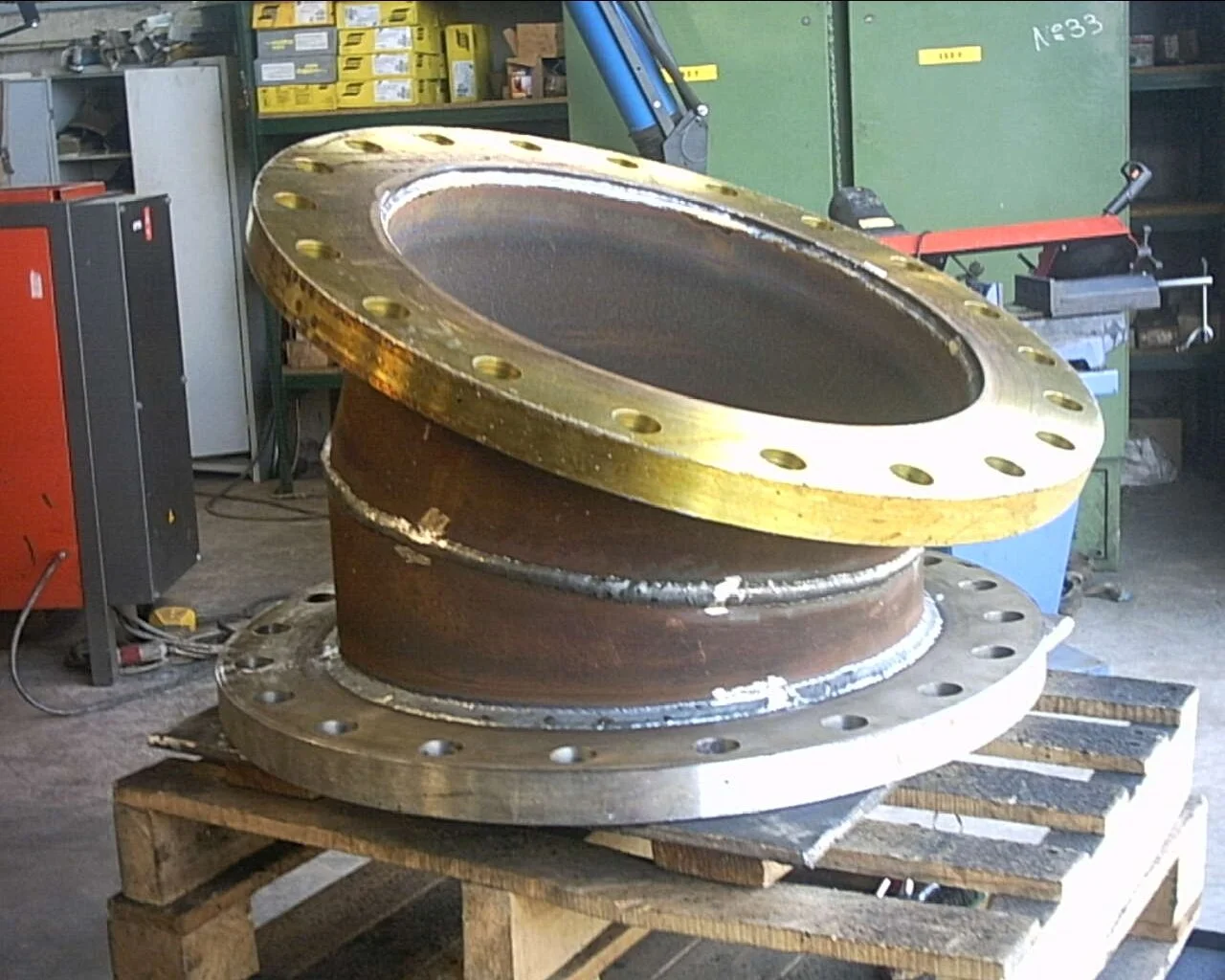 Componente industriale di grandi dimensioni con flange metalliche, appoggiato su un pallet di legno, in un'officina.