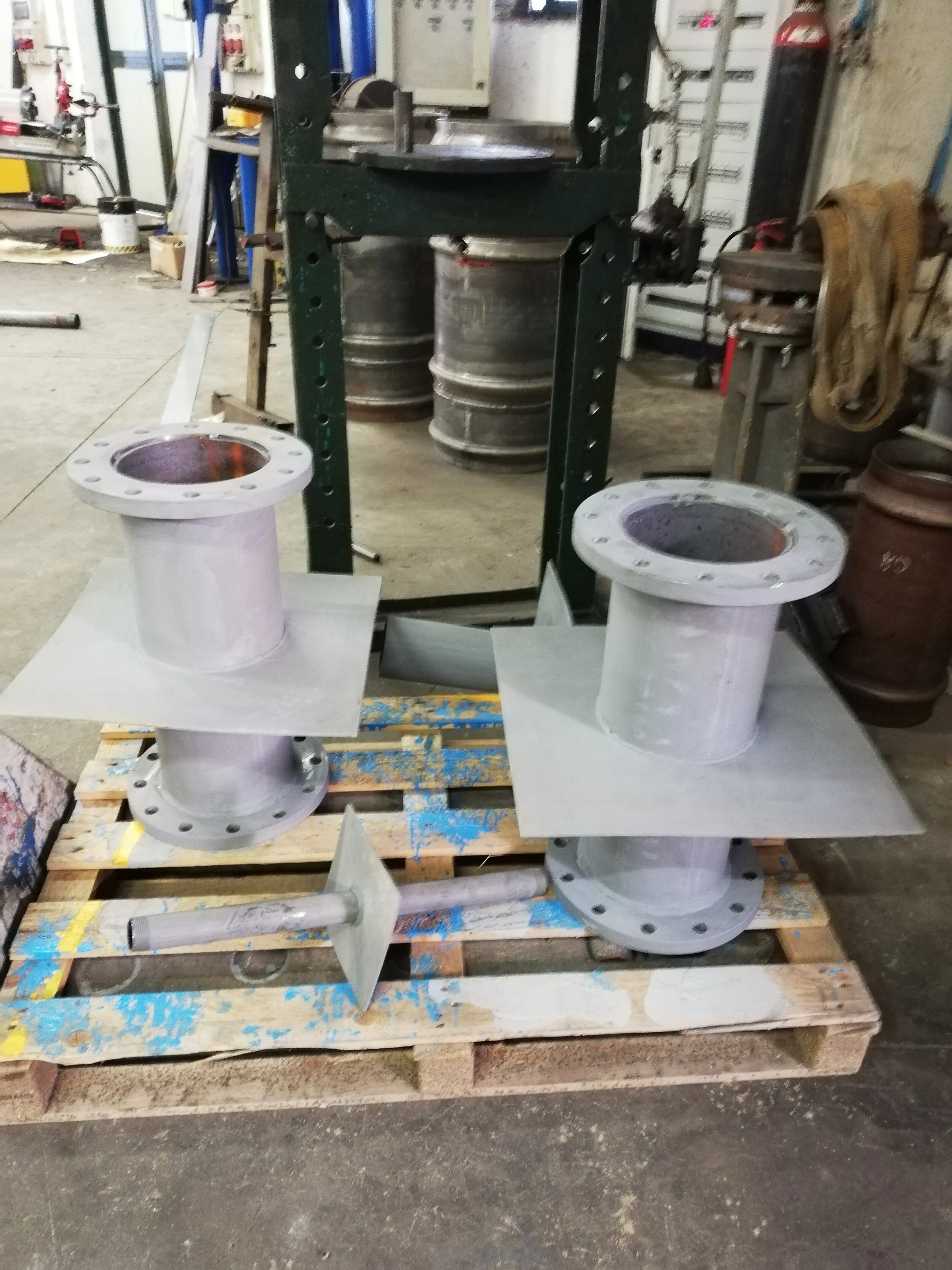 Due componenti industriali in metallo con flange, posizionati su una pedana di legno in un'officina.