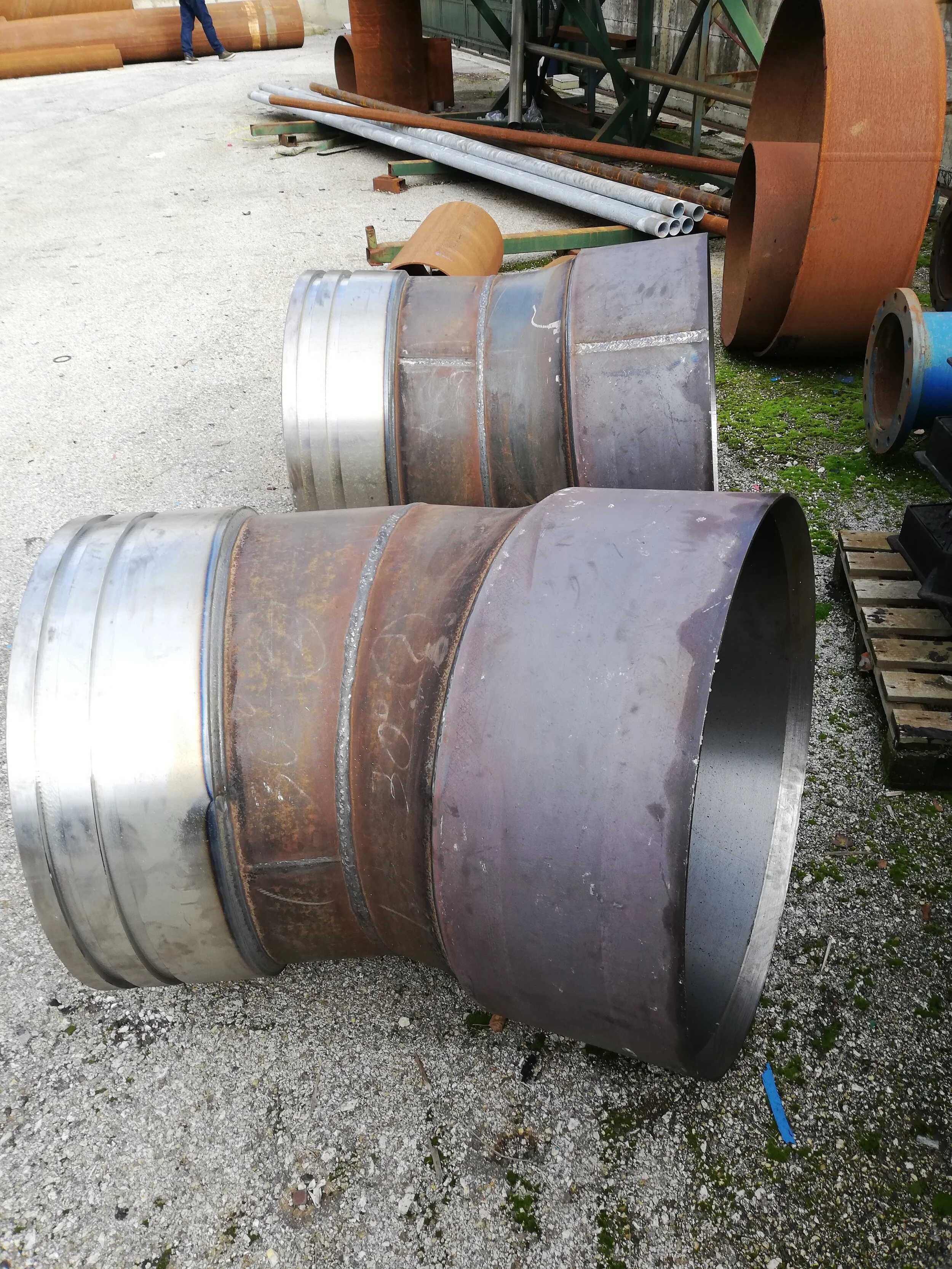 Due grandi pezzi di metallo cilindrici, probabilmente parti di tubi o contenitori industriali, giacciono sul suolo in un'area di lavoro con altri tubi e attrezzature in background.