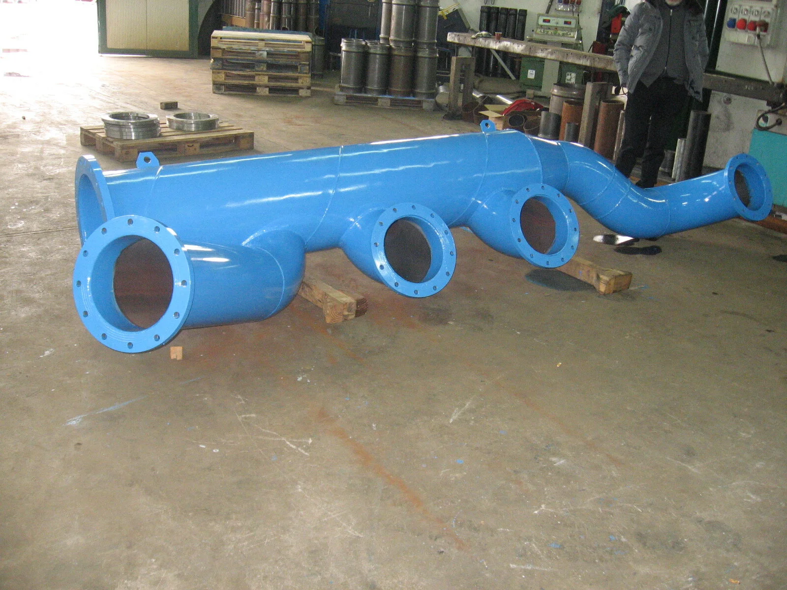 Un grande tubo blu con più aperture, posizionato su un pavimento industriale, in un'officina o fabbrica.