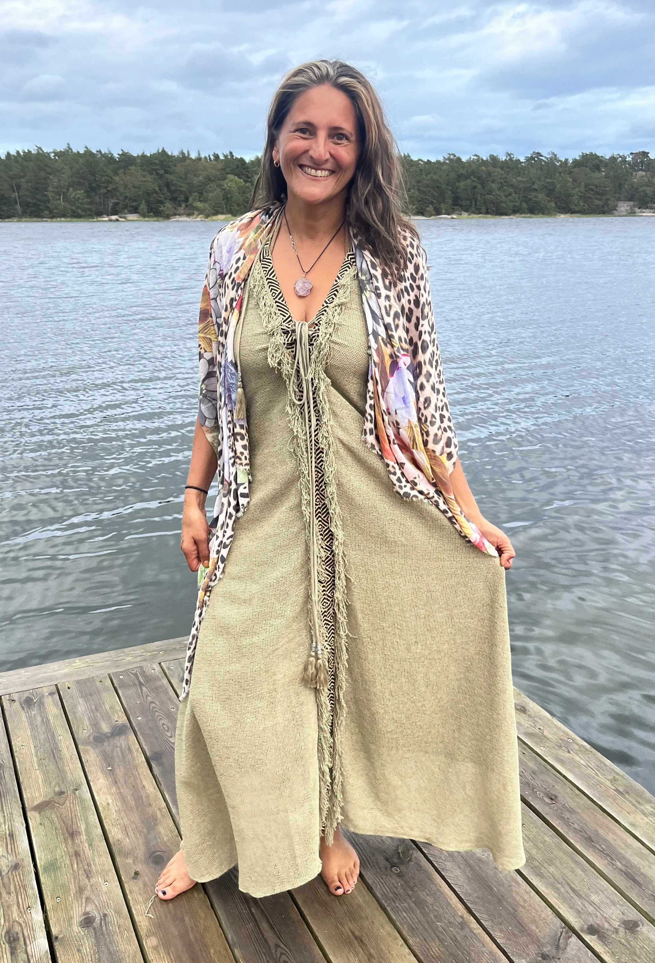 A woman standing on a wooden dock by a lake with a forested shoreline behind her. The woman has long, wavy hair and is smiling. She is wearing a beige dress and an open, patterned kimono-style cover-up. She is barefoot, and the sky is cloudy.