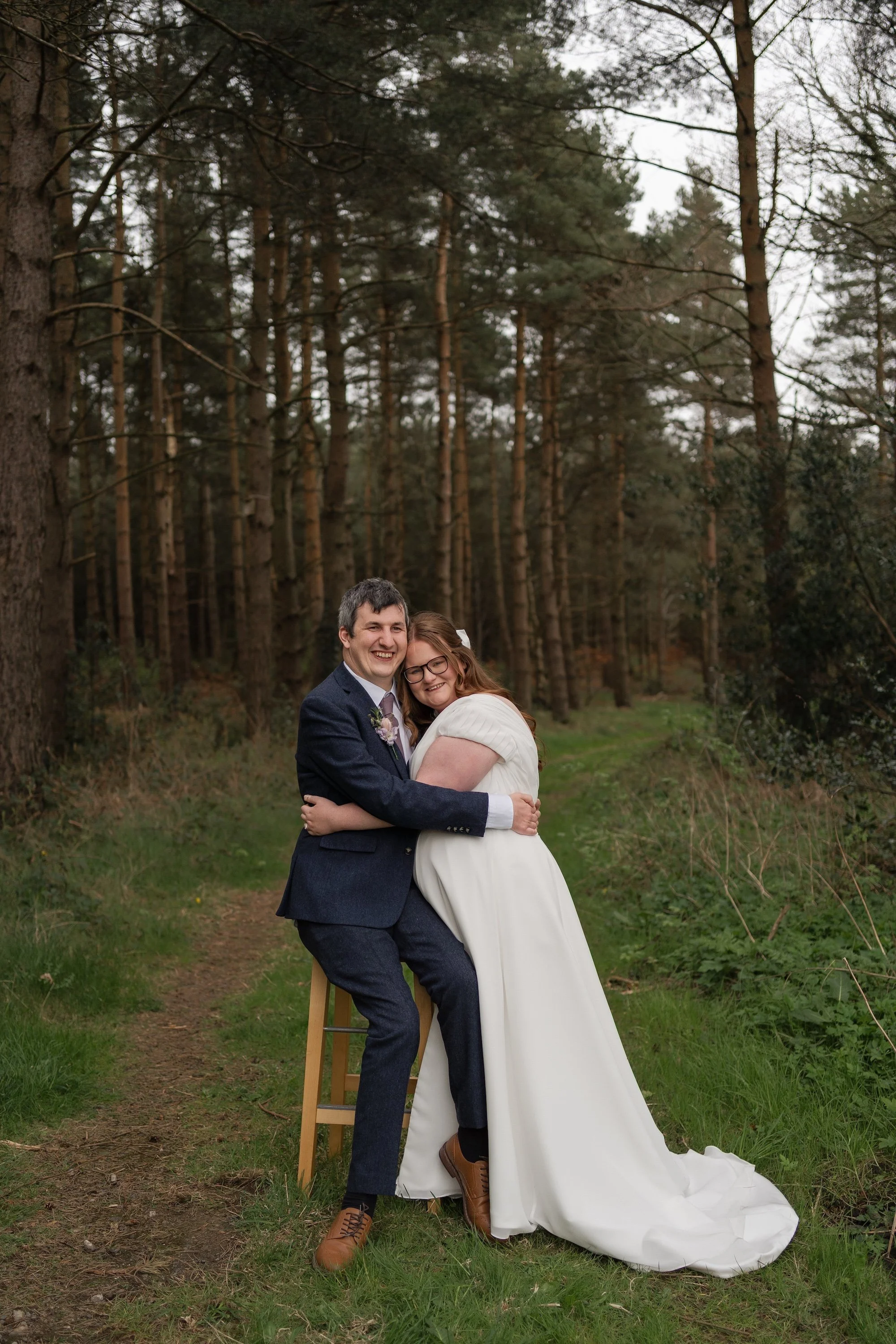 A couple, dressed in wedding attire, embracing each other in a forest with tall trees and green grass at Healey Barn.