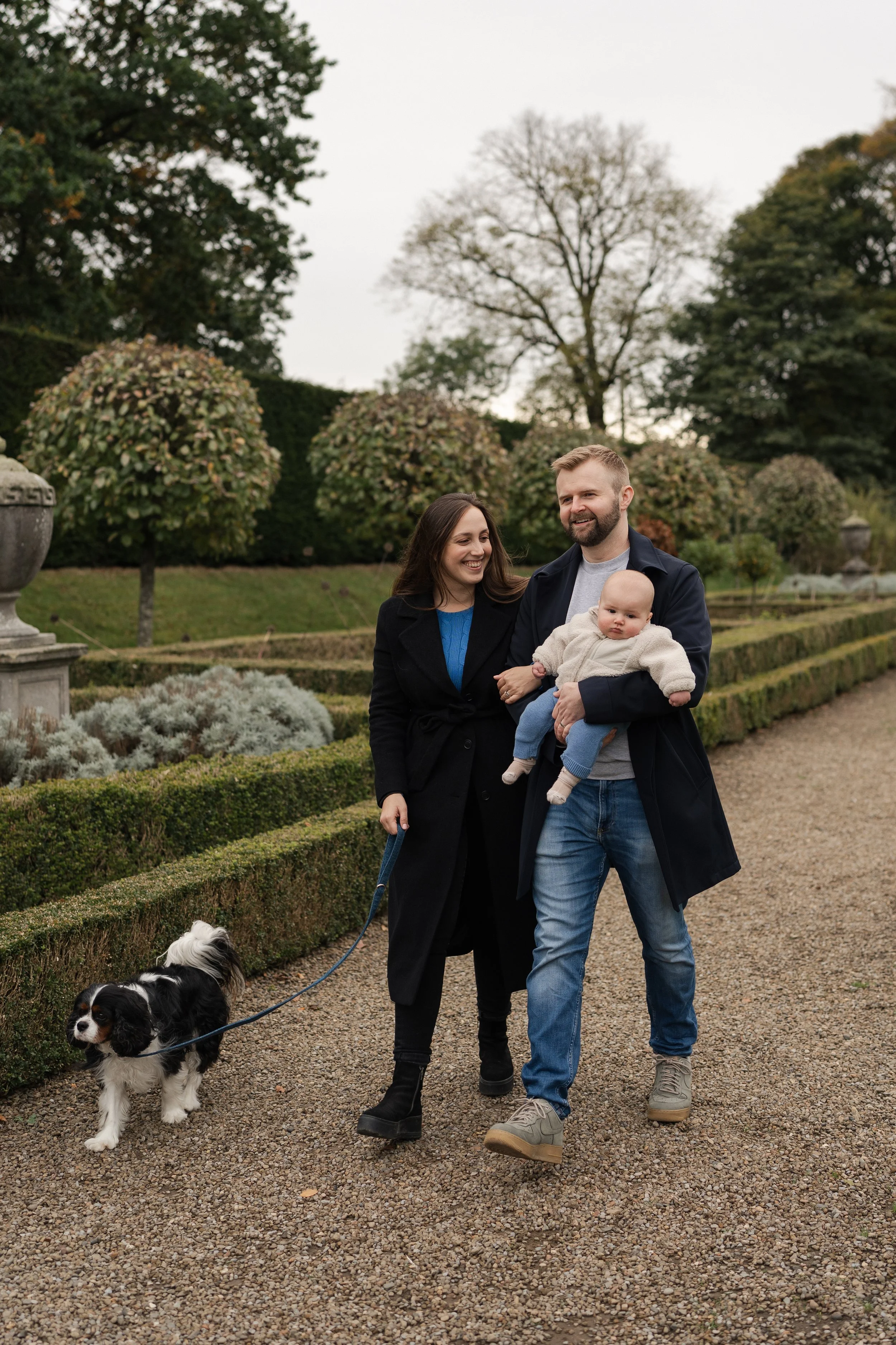 A family of three walking their dog in a garden during daytime. The woman and man are smiling while holding a baby, and the dog is on a leash.