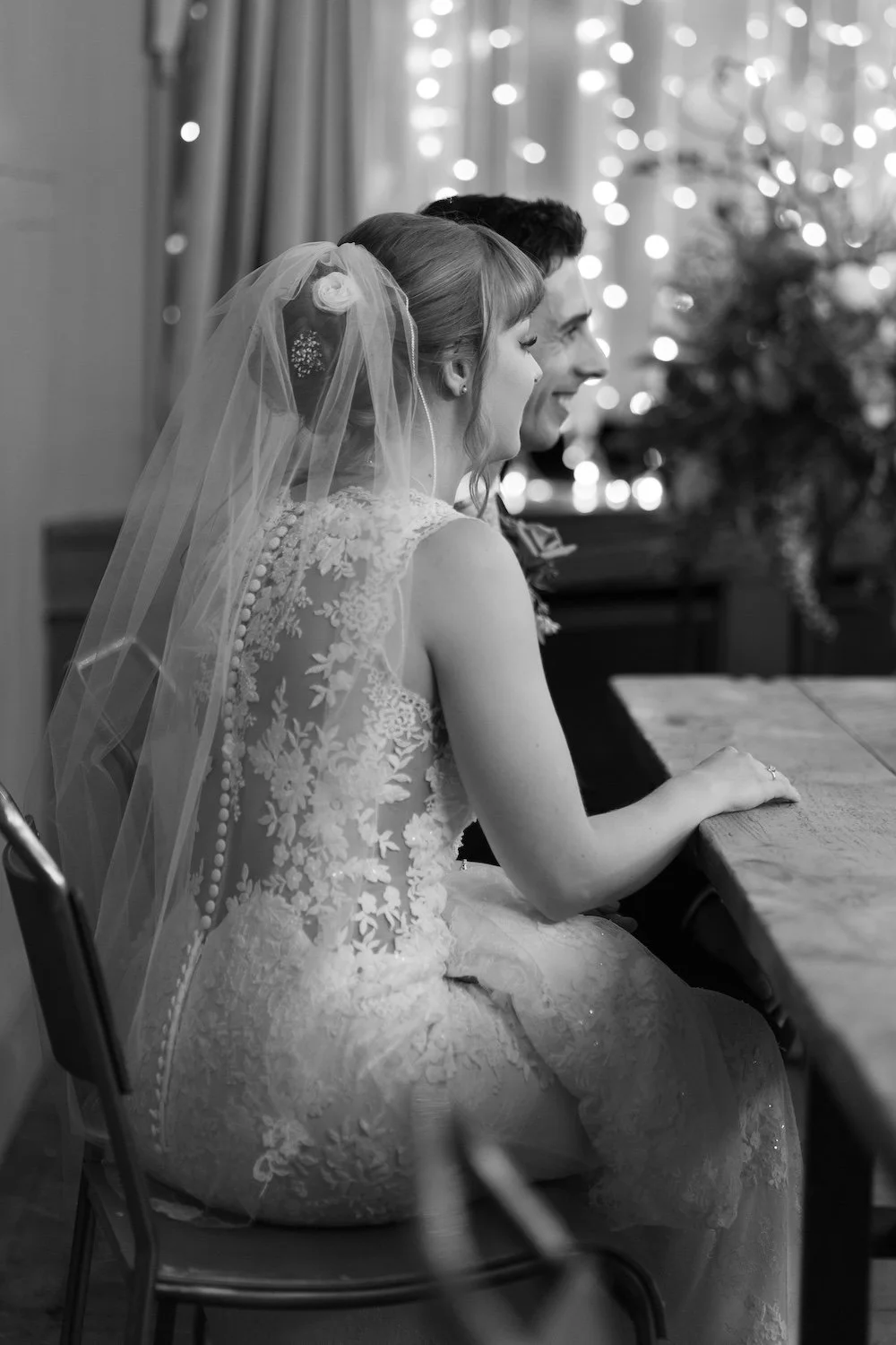 A bride and groom sitting side by side at a wedding reception, smiling, with festive lights in the background. Wylam Brewery.