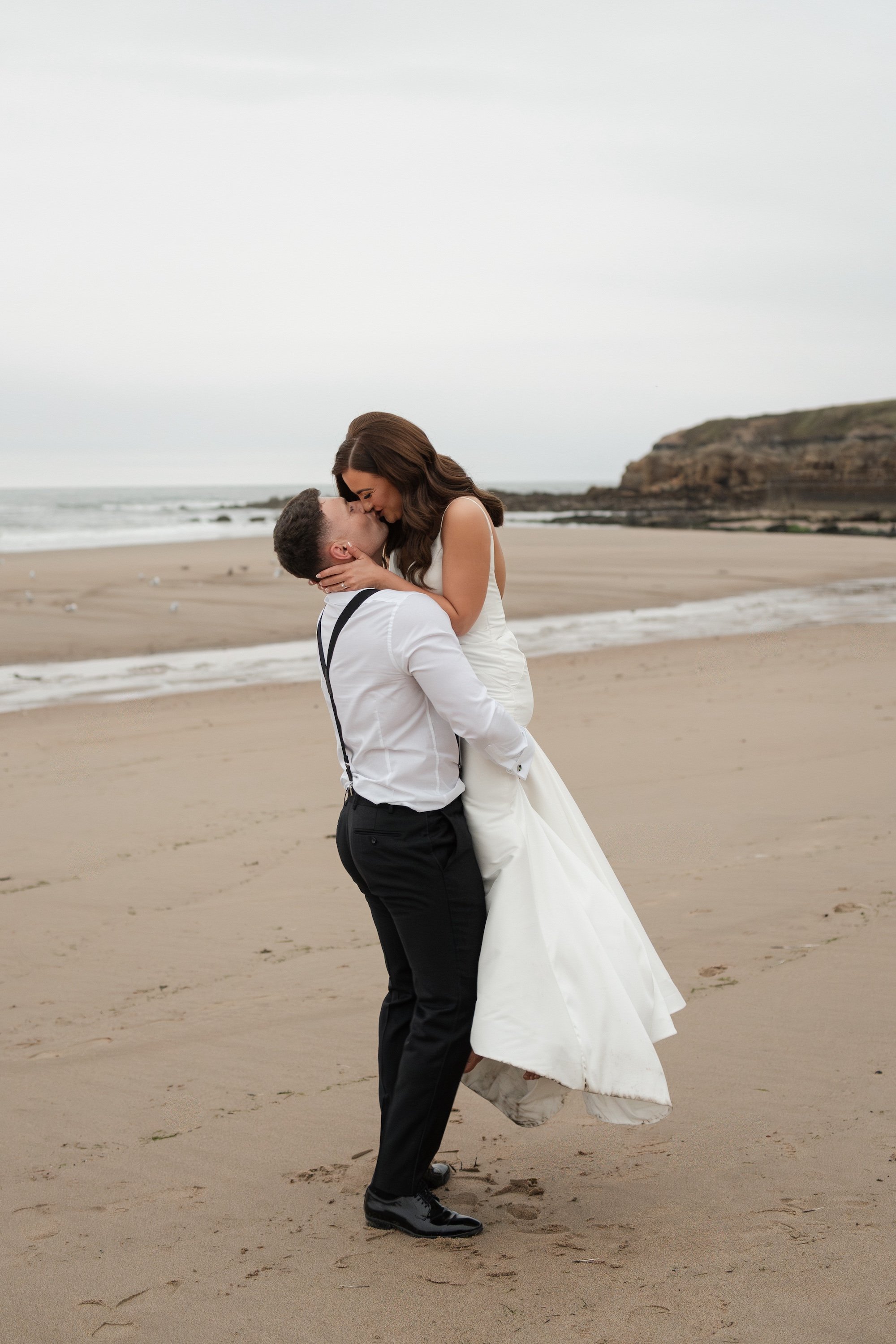 A couple in wedding attire sharing a kiss on the beach, with the groom lifting the bride. Tynemouth Beach .
