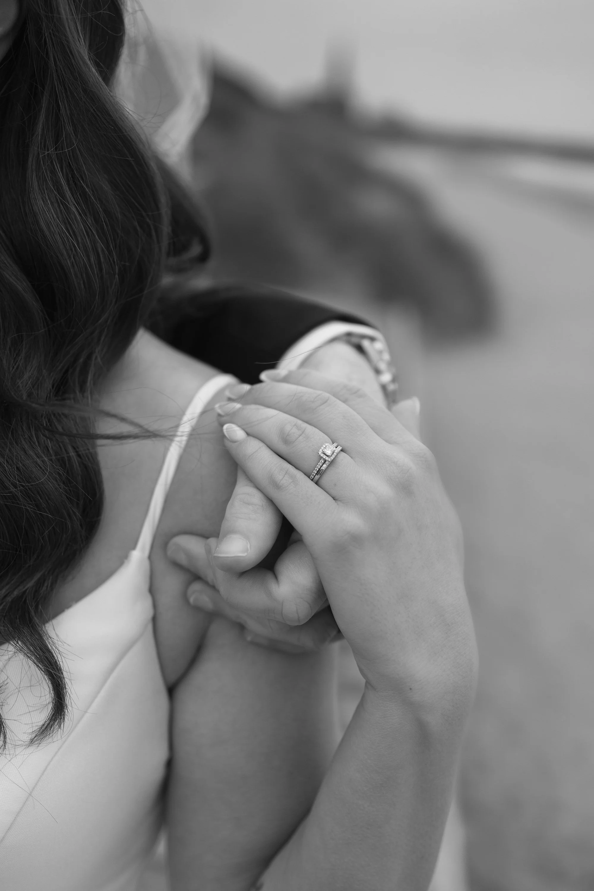 Close-up of a woman with long wavy hair wearing a wedding ring on her finger, touching her shoulder in black and white.