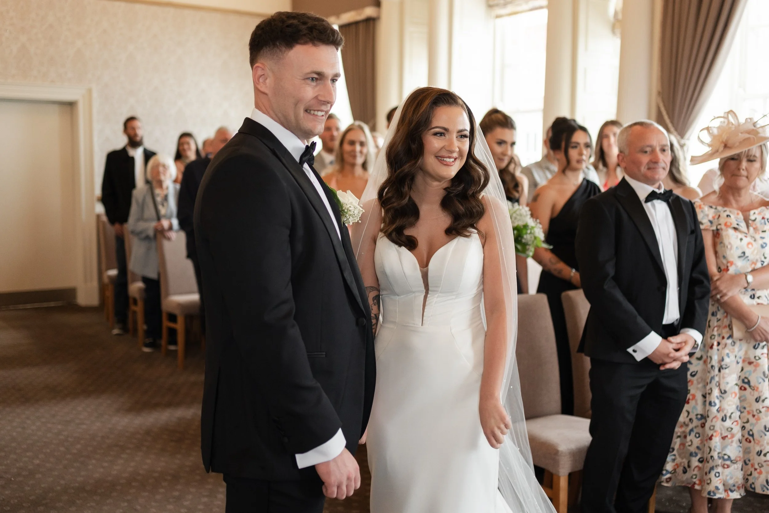 A bride and groom standing and smiling during their wedding ceremony with family and friends in the background. North Tyneside Registry Office.