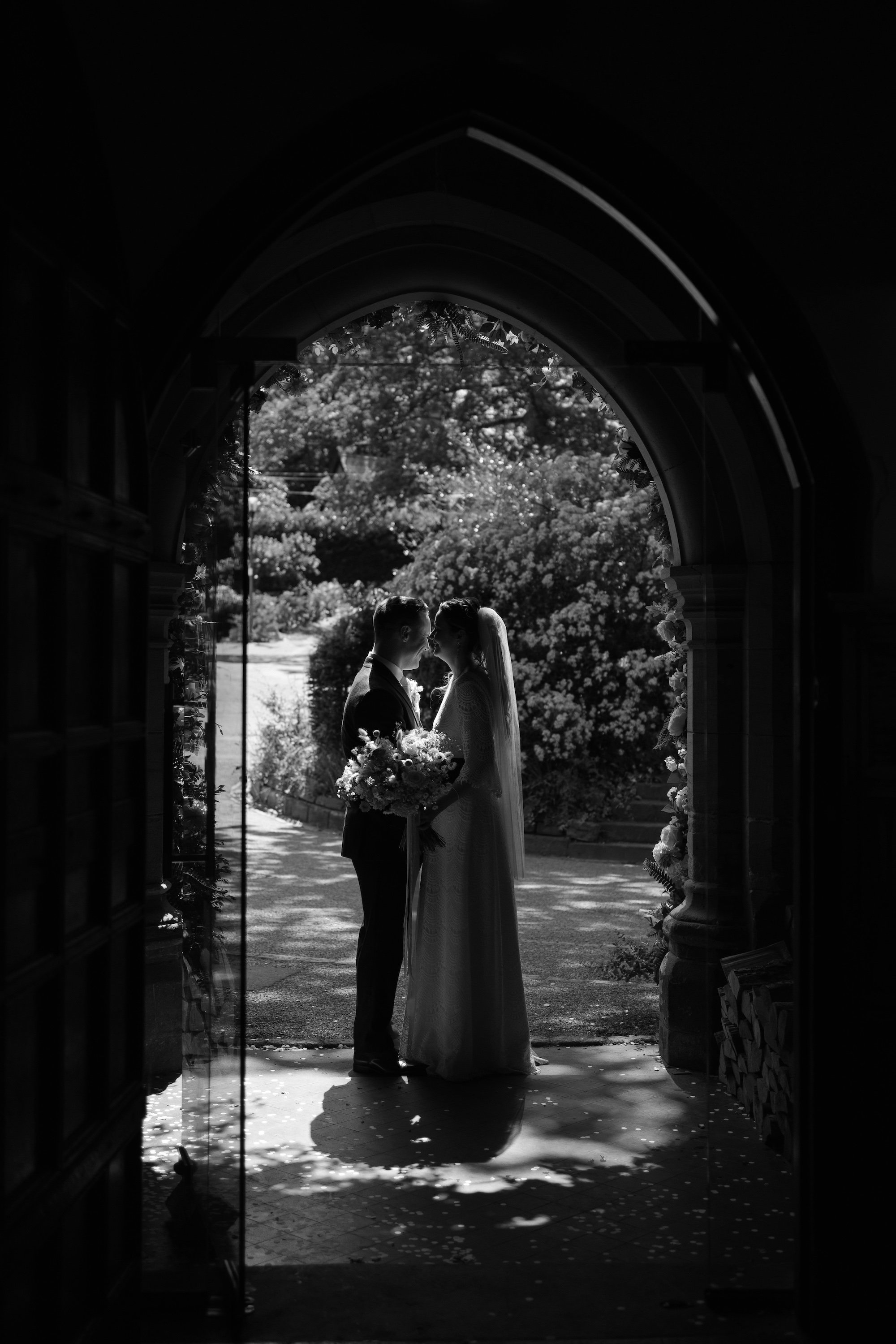 A black and white photo of a bride and groom standing close together, holding a bouquet, seen through an arched doorway decorated with flowers, with a garden in the background. Jesmond Dene House. 