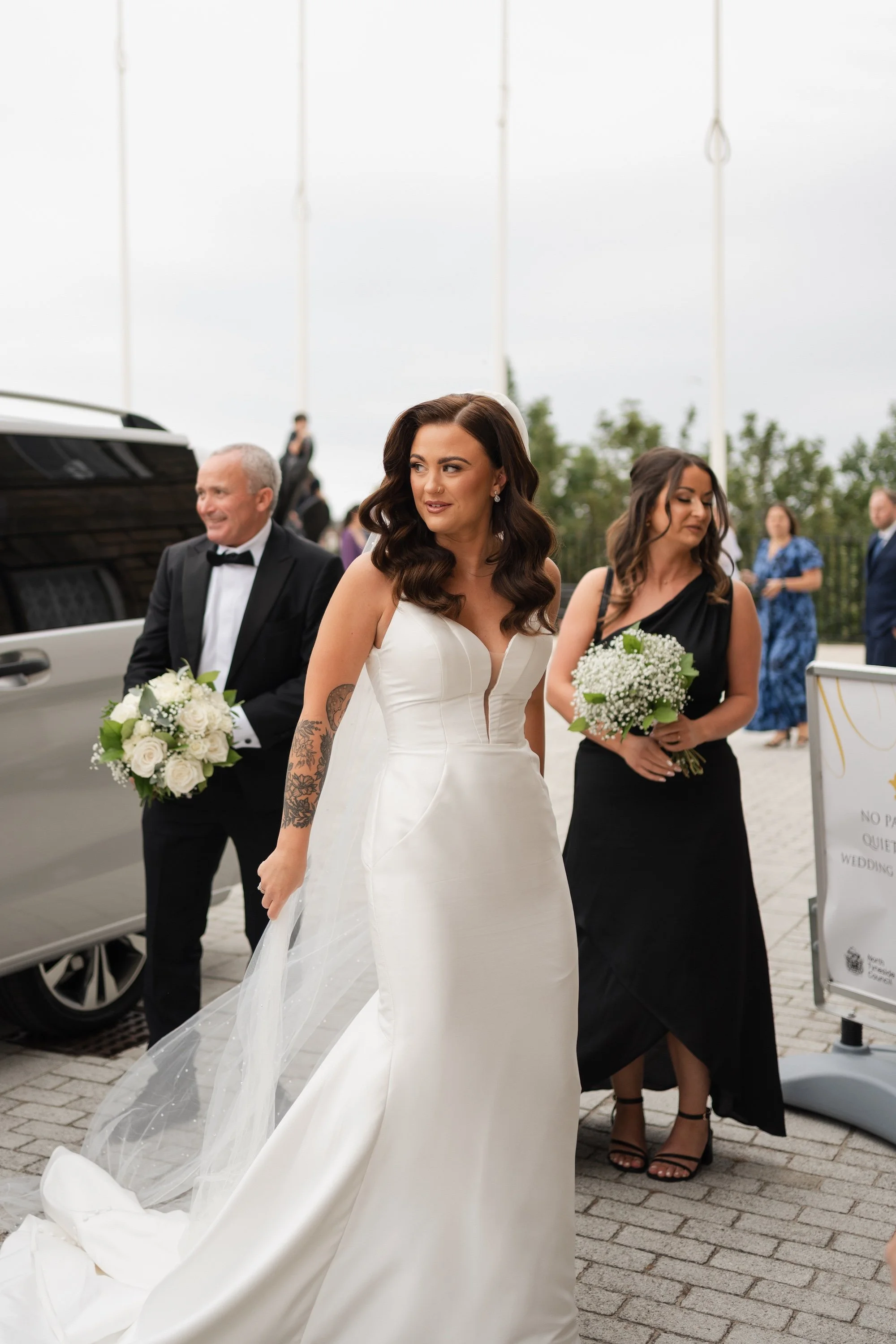 Bride in a white wedding dress with dark wavy hair, standing outdoors near a limousine, surrounded by bridesmaids and guests. Bride has a tattoo on her left arm and is holding her train. North Shields Registry Office.