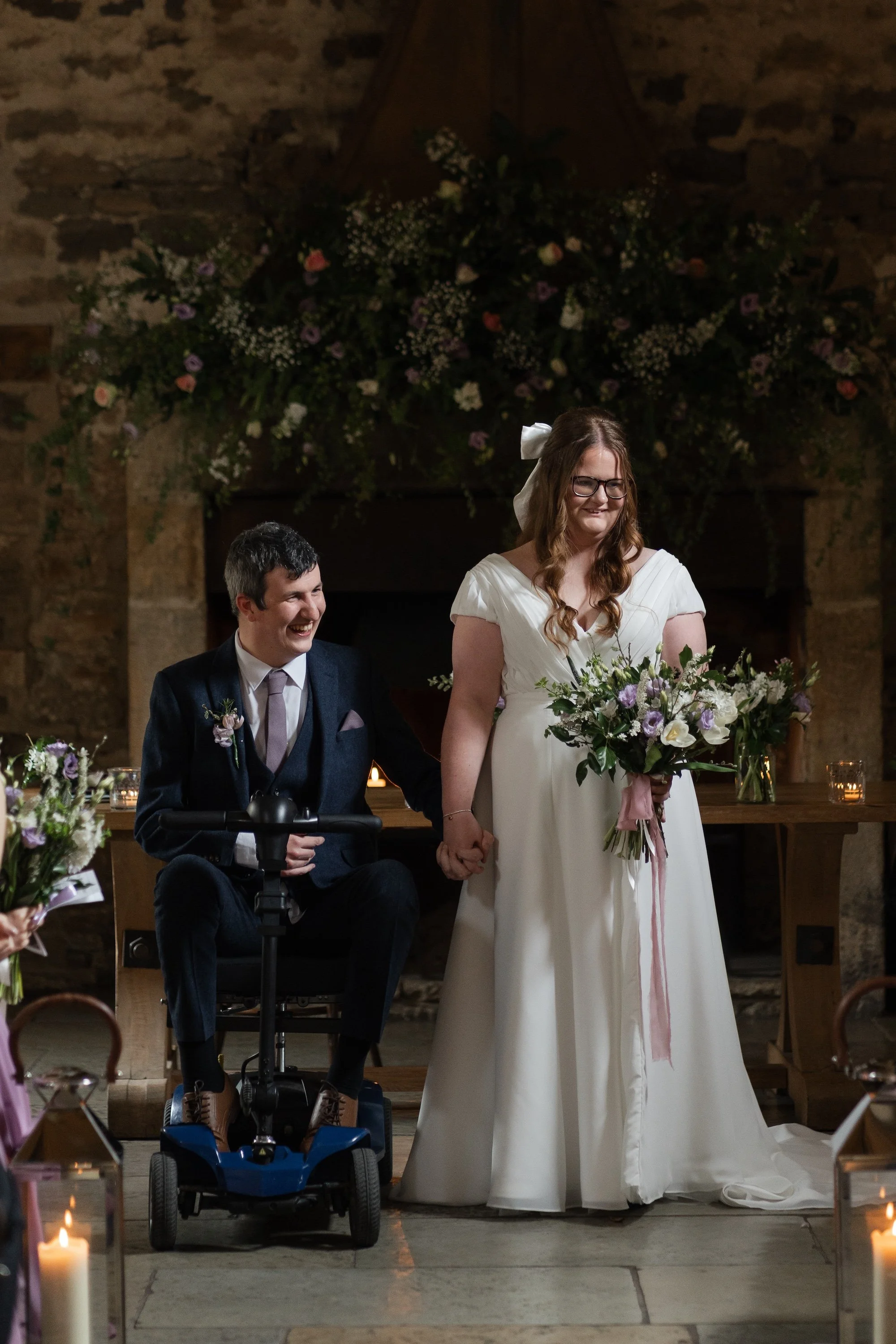 A bride and groom holding hands during their wedding ceremony, with the groom in a wheelchair. Healey Barn. 