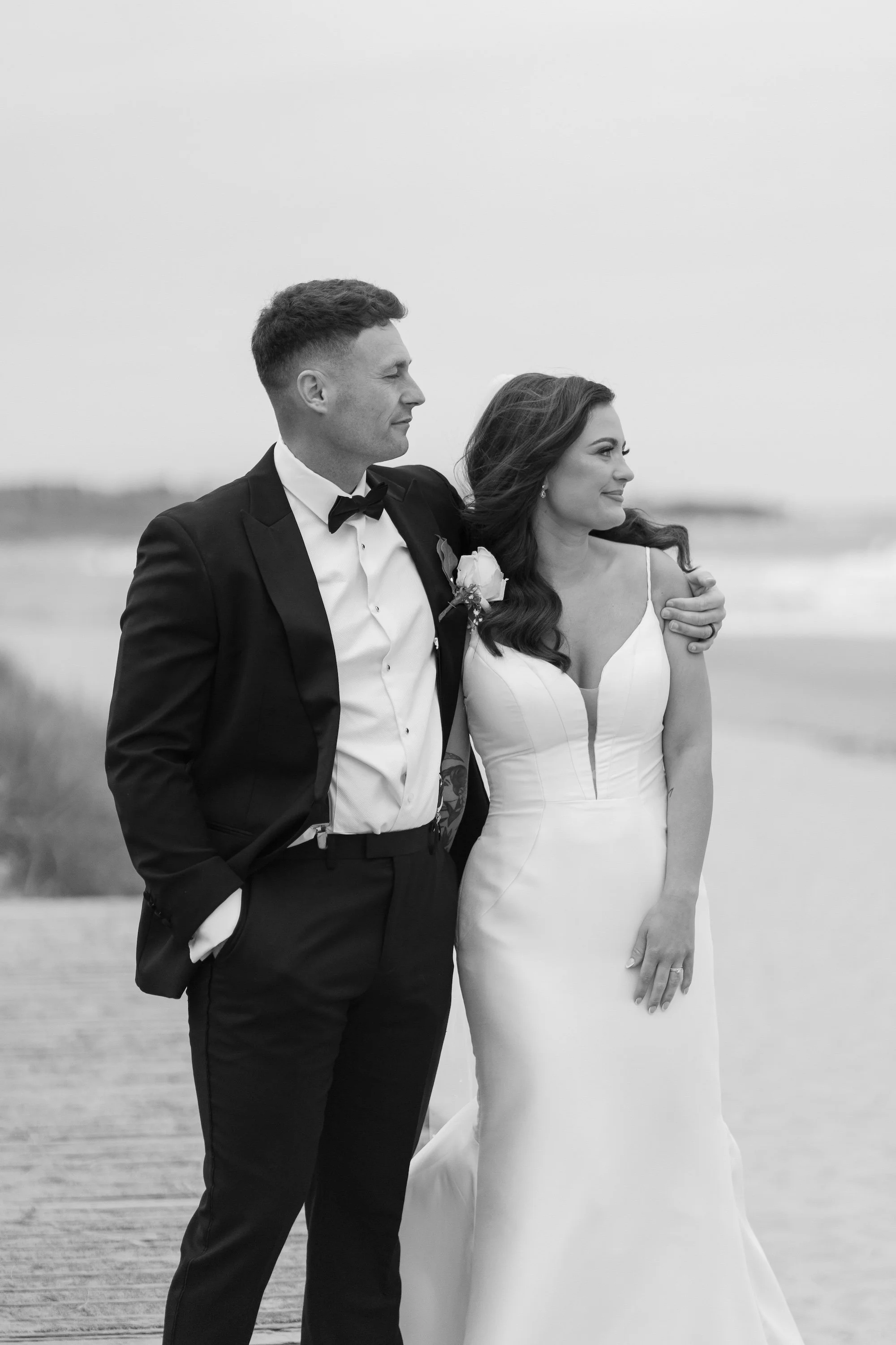 A black-and-white photograph of a bride and groom dressed in wedding attire on a beach, with the groom in a tuxedo and the bride in a white gown, standing close and looking into the distance.