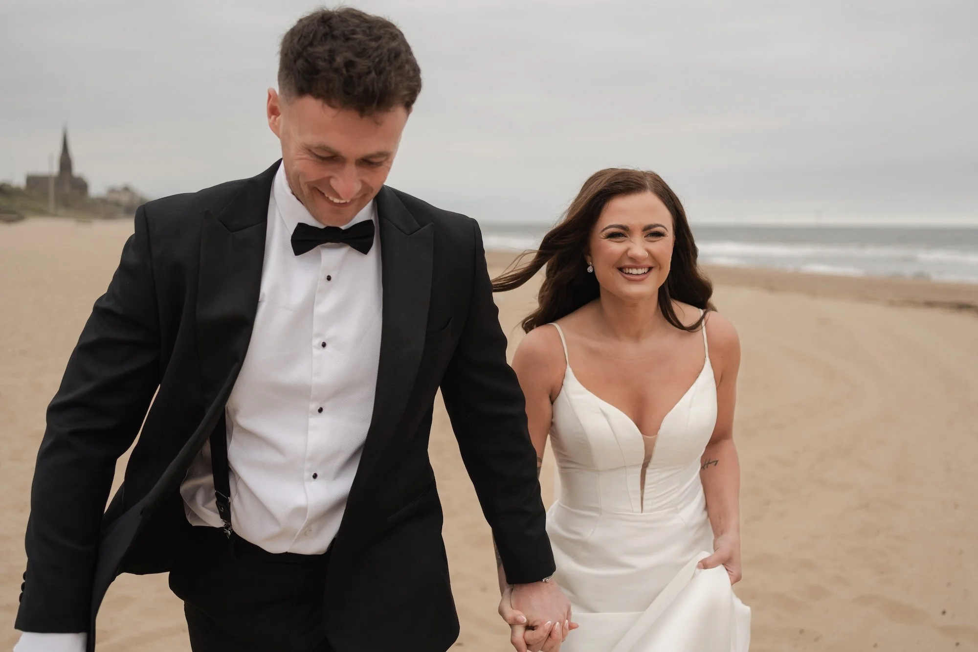 A newlywed couple dressed in wedding attire walking on the beach, holding hands and smiling. Longsands, Tynemouth. 