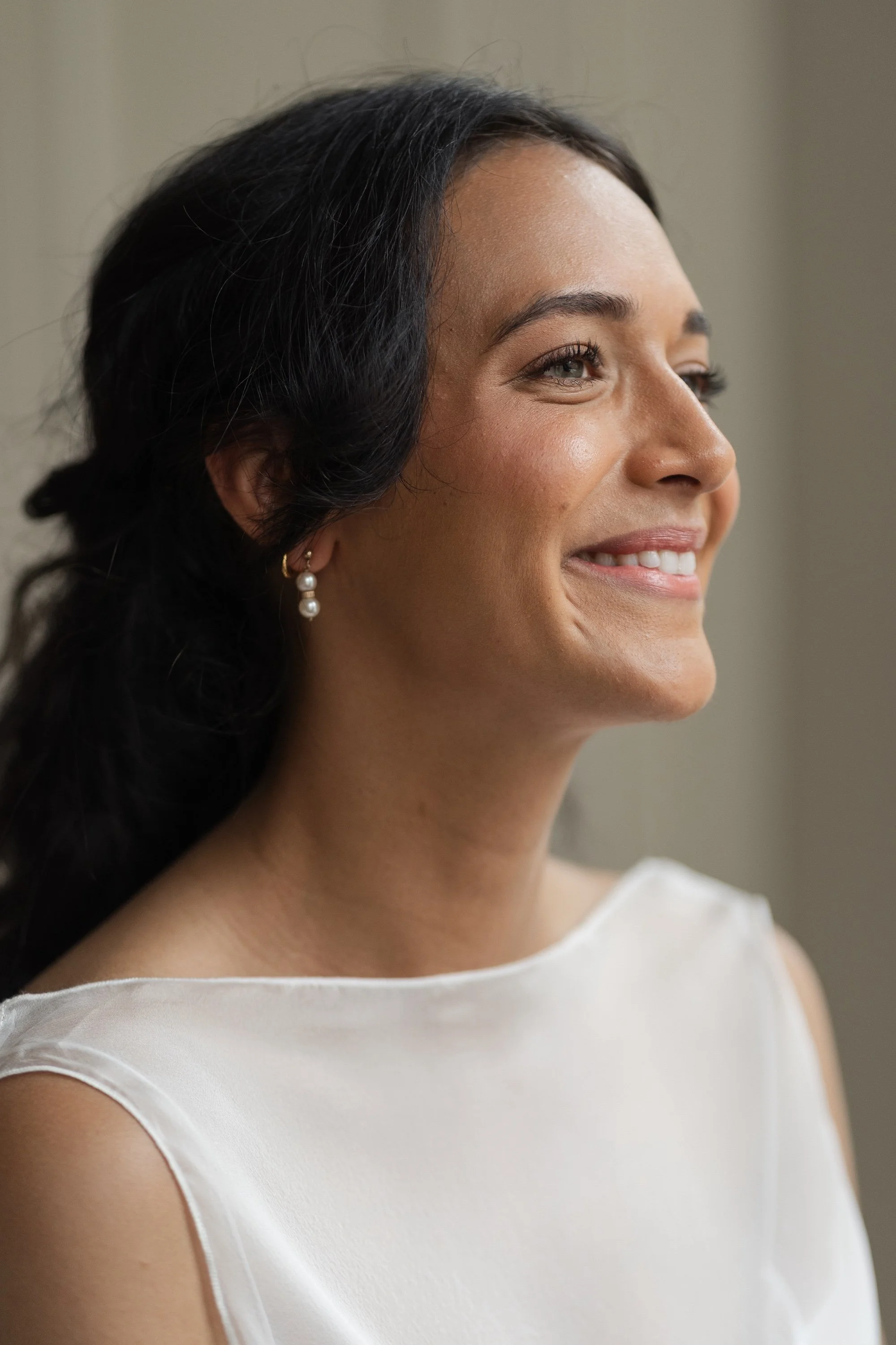 A woman with dark hair and pearl earrings smiling, wearing a white sleeveless top.