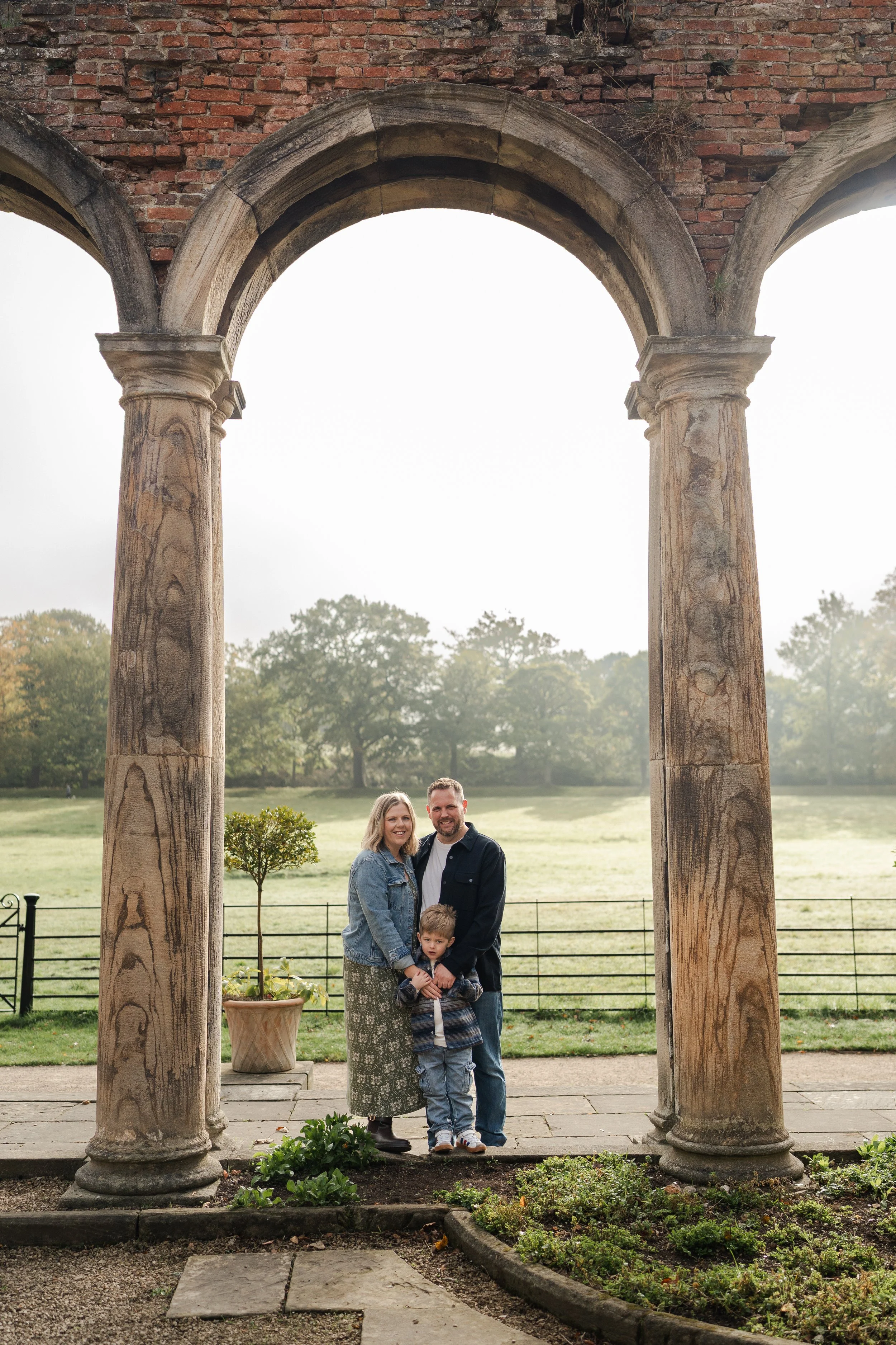 A family of three standing together under ancient stone columns with a scenic green field and trees in the background. North Tyneside family photos. 