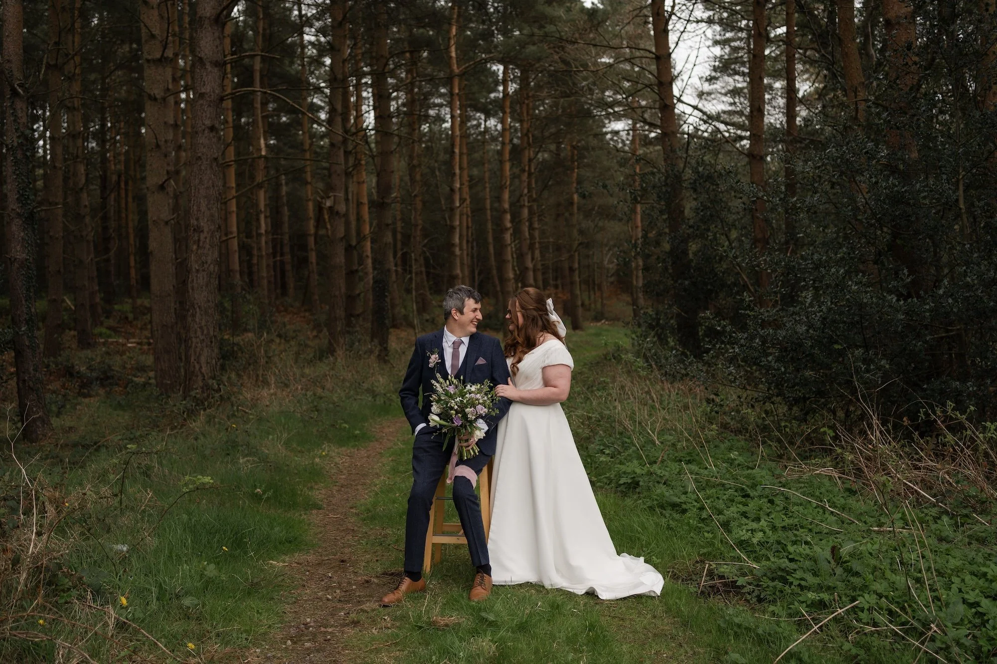 A bride and groom standing on a forest trail, smiling at each other. The groom is dressed in a dark suit, and the bride is in a white wedding dress with a bow in her hair. The groom holds a bouquet of flowers. Healey Barn. 