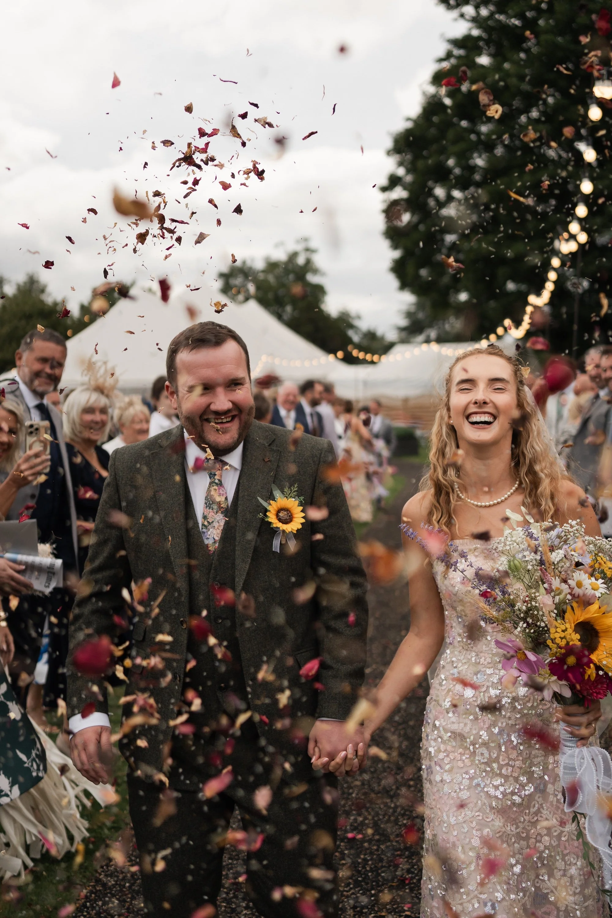 A couple is smiling and holding hands during their wedding celebration, surrounded by guests throwing flower petals in an outdoor setting with string lights and a white tent in the background.