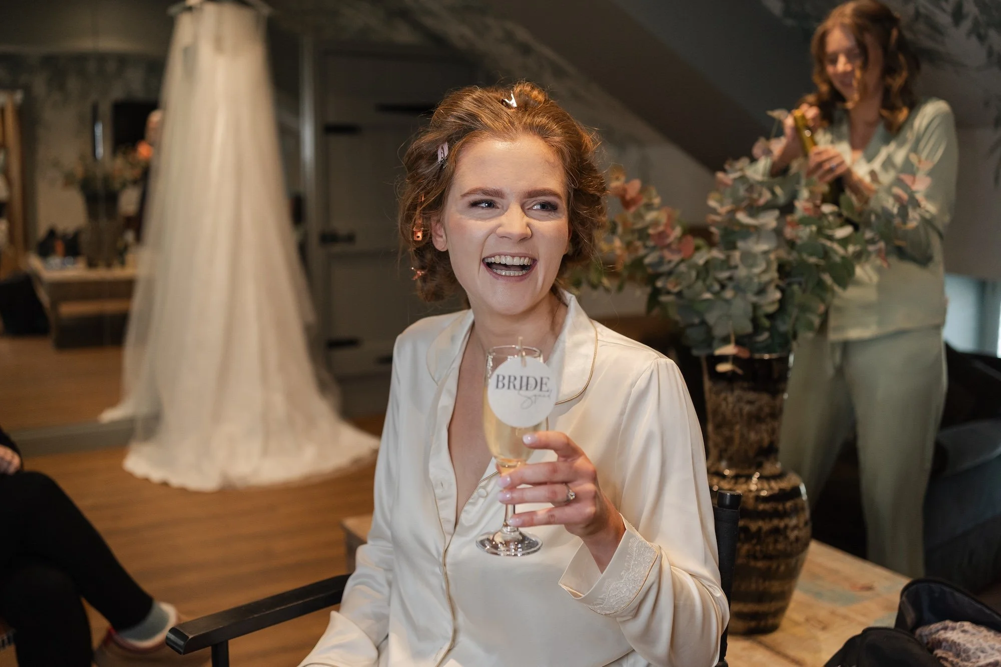 A woman in satin pajamas sitting indoors, holding a glass of champagne with a 'Bride' tag, smiling and making a face, with another woman in pajamas behind her holding a bouquet of flowers.