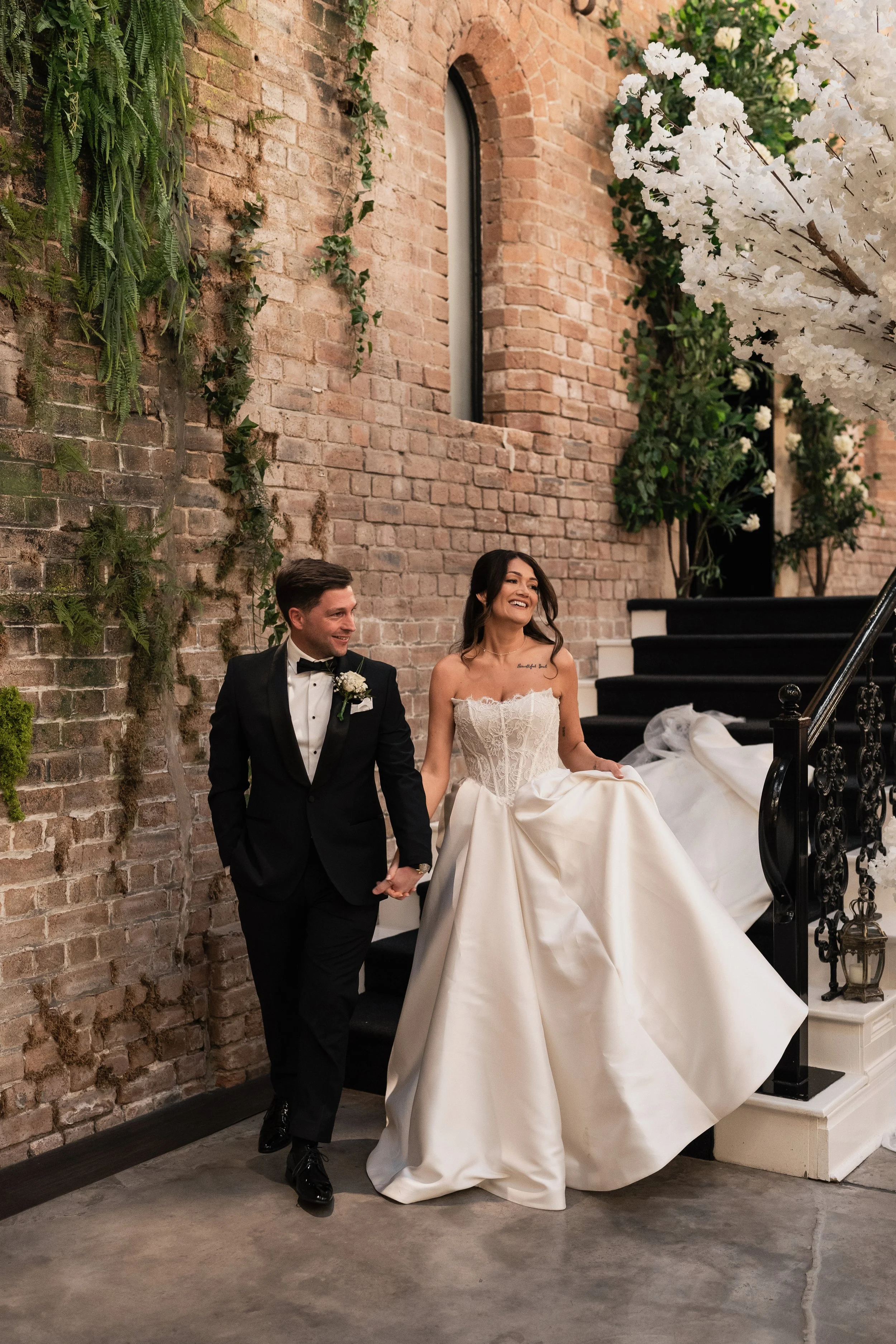 A bride and groom holding hands and walking down stairs inside a rustic brick venue, surrounded by green plants and white flowers. Dalton Old Pump House. 