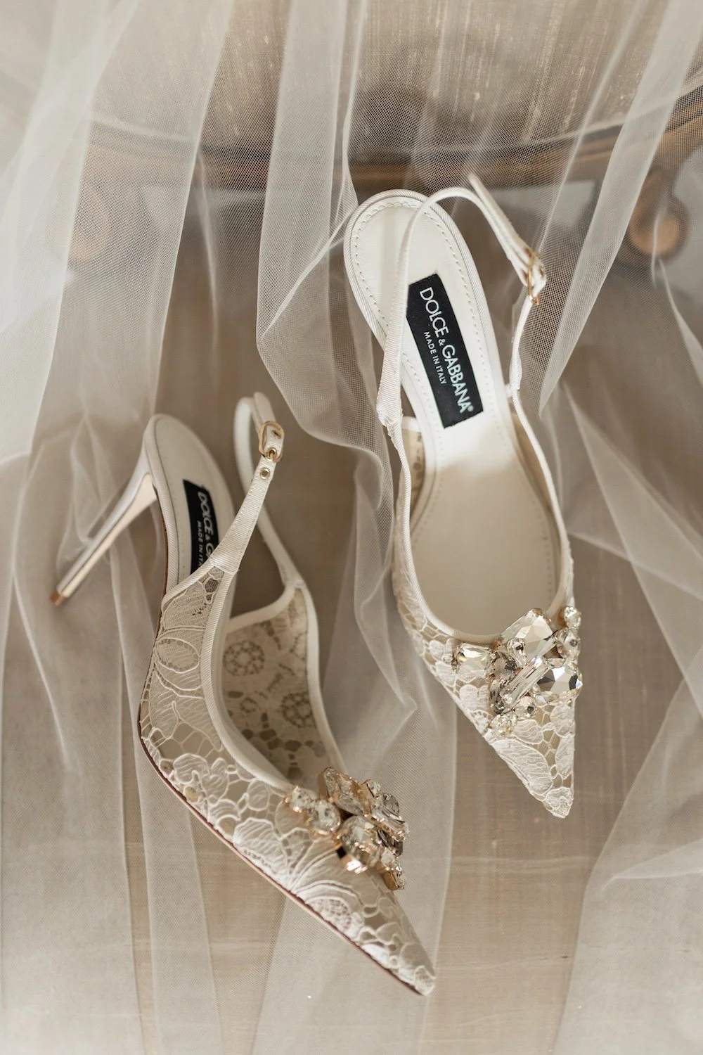 Elegant white lace high-heeled shoes with large rhinestone embellishments on the pointed toes, placed on sheer fabric.
