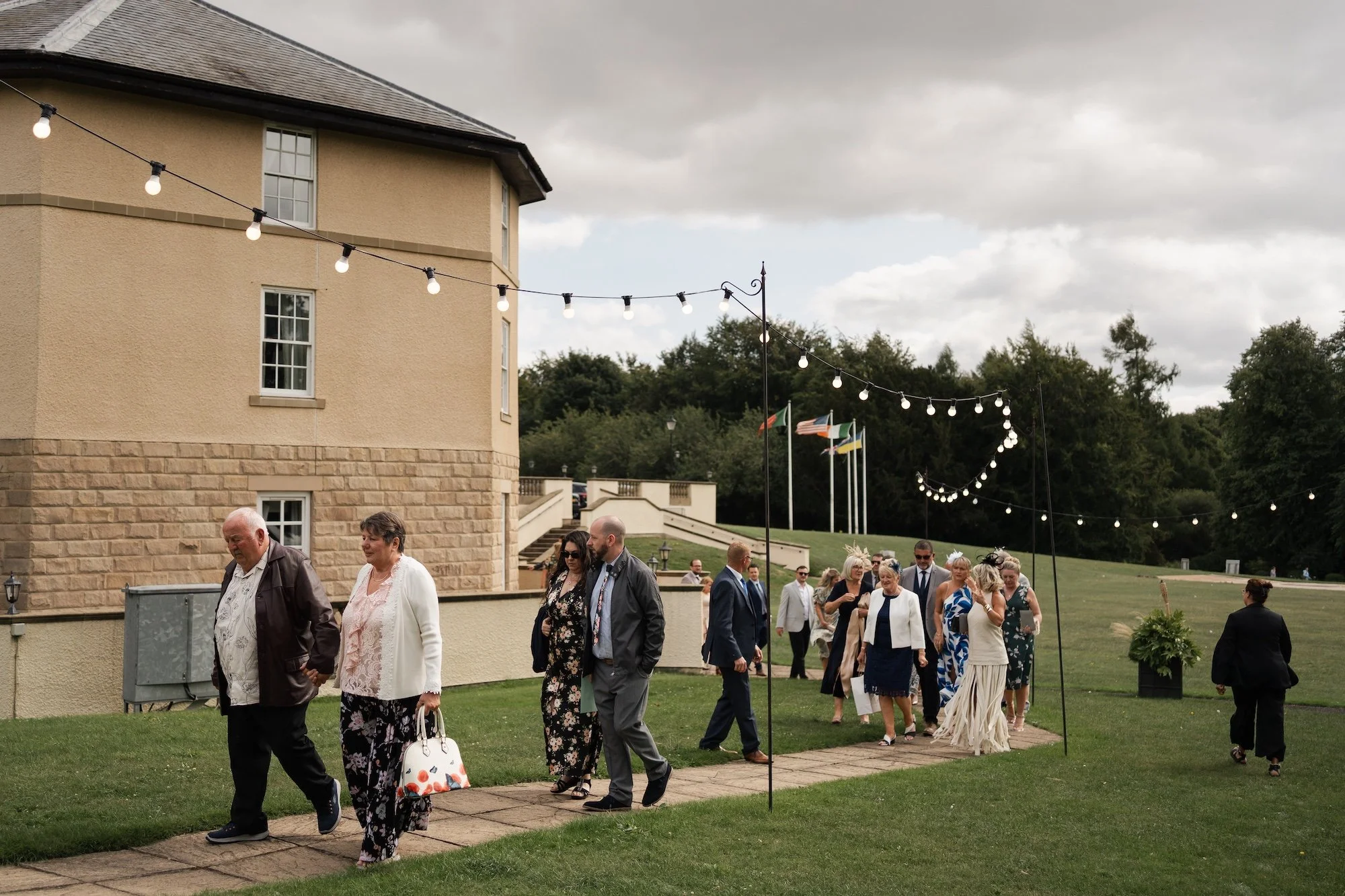 People dressed in formal attire walking along a brick pathway outdoors under string lights, near a beige building with flags in the background on a cloudy day. Hardwick Hall Hotel. 