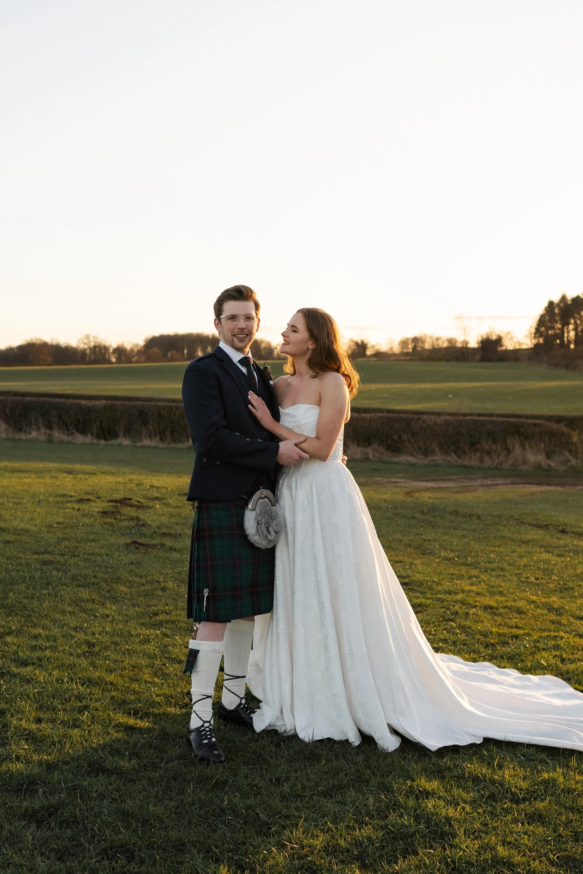 A bride and groom stand together on a grassy field during sunset, with the bride wearing a white wedding dress and the groom wearing a kilt and jacket. Doxford Barns.
