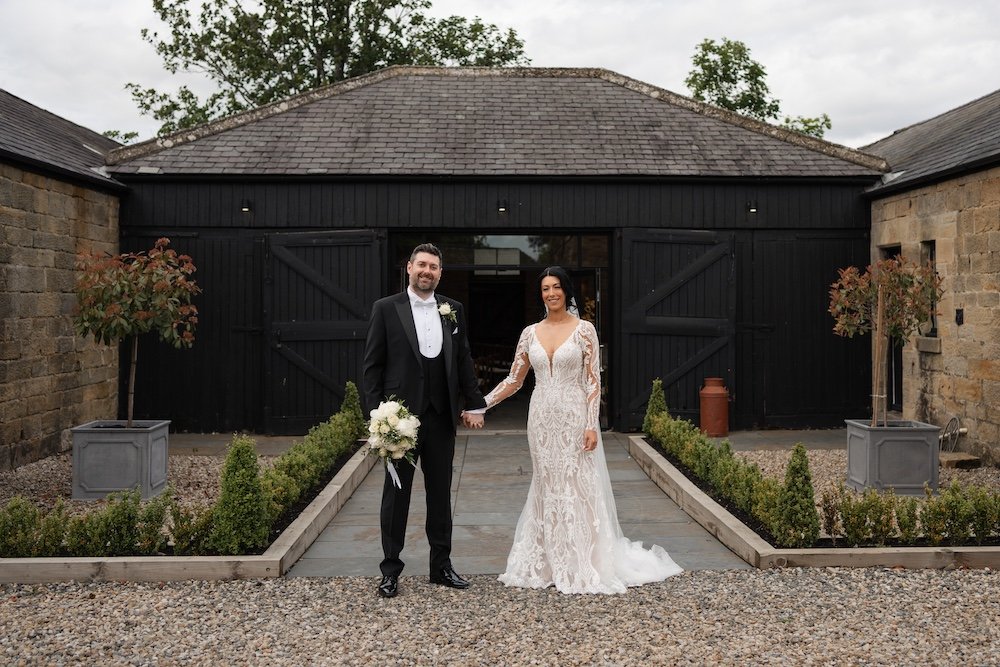 A bride and groom holding hands in front of a black barn at a wedding venue, with potted trees and small bushes along the walkway. Horton Grange.