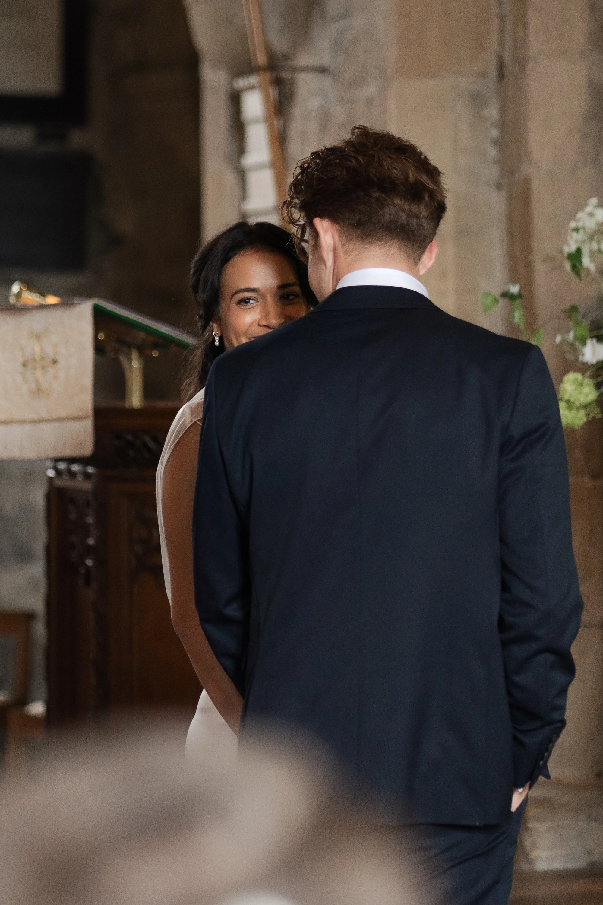 A bride and groom standing close together during their wedding ceremony, with the bride smiling and looking at the groom, who has his back to the camera.