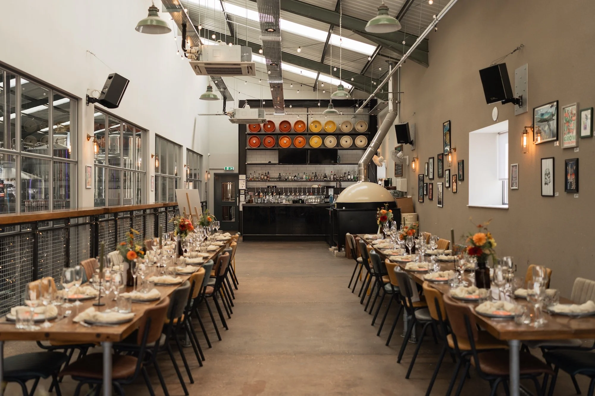 An elegant event space with two long wooden tables set with white napkins, glassware, and floral centerpieces, arranged for a celebration or gathering. Full Circle Brewery wedding reception. 