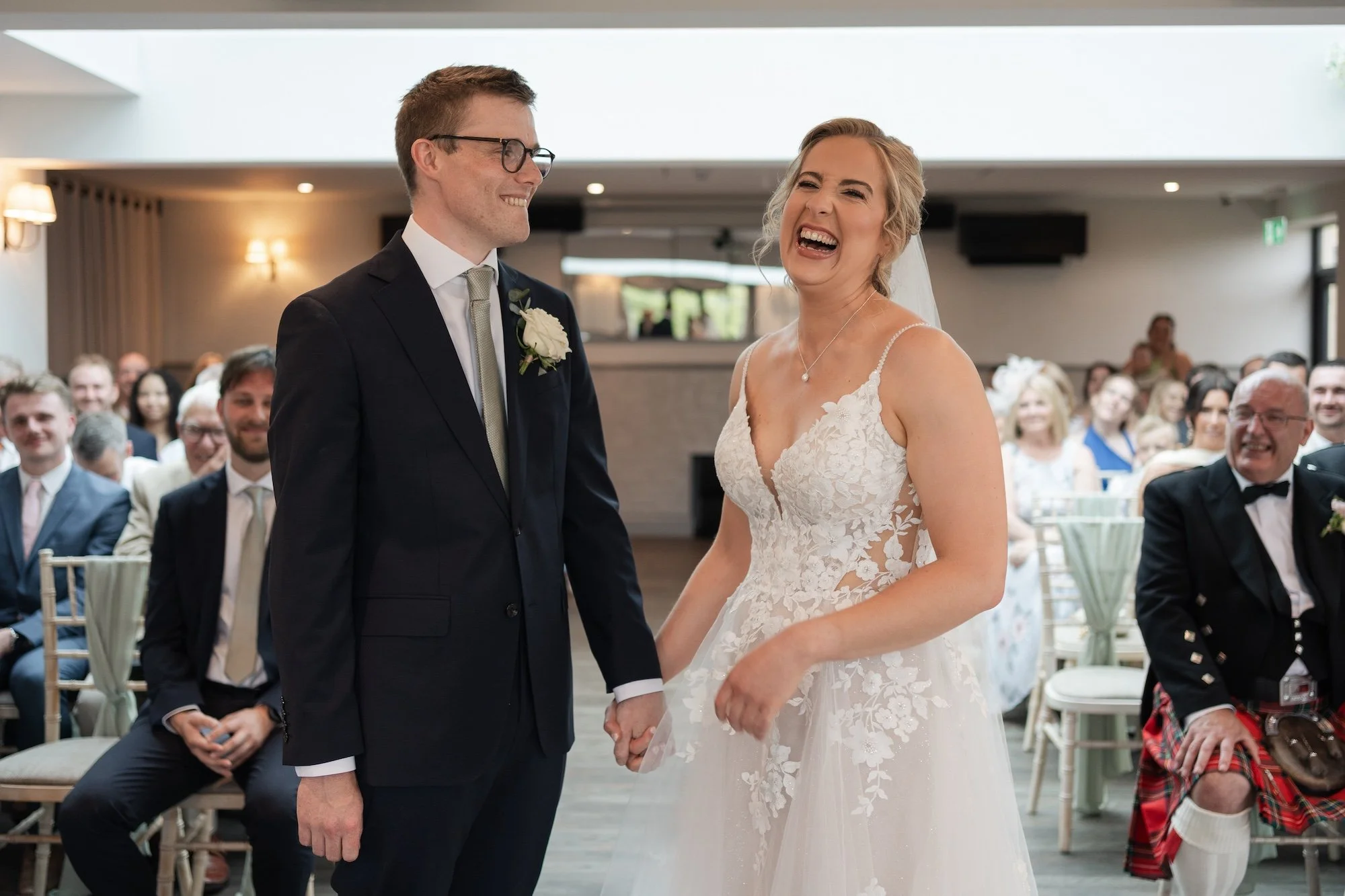 A bride and groom holding hands during their wedding ceremony, surrounded by seated guests, in an indoor venue with natural and warm lighting. South Causey Inn.