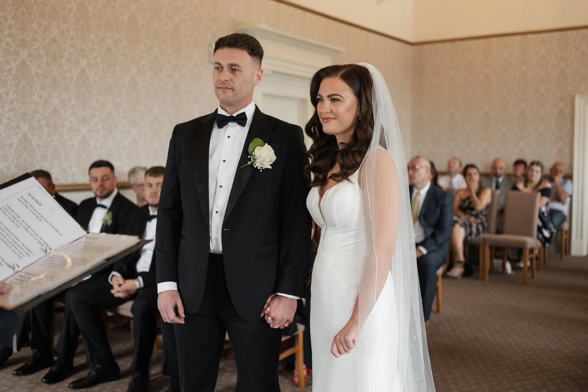 A bride and groom standing hand-in-hand during their wedding ceremony in a church, with guests seated in the background. North Tyneside registry office. 